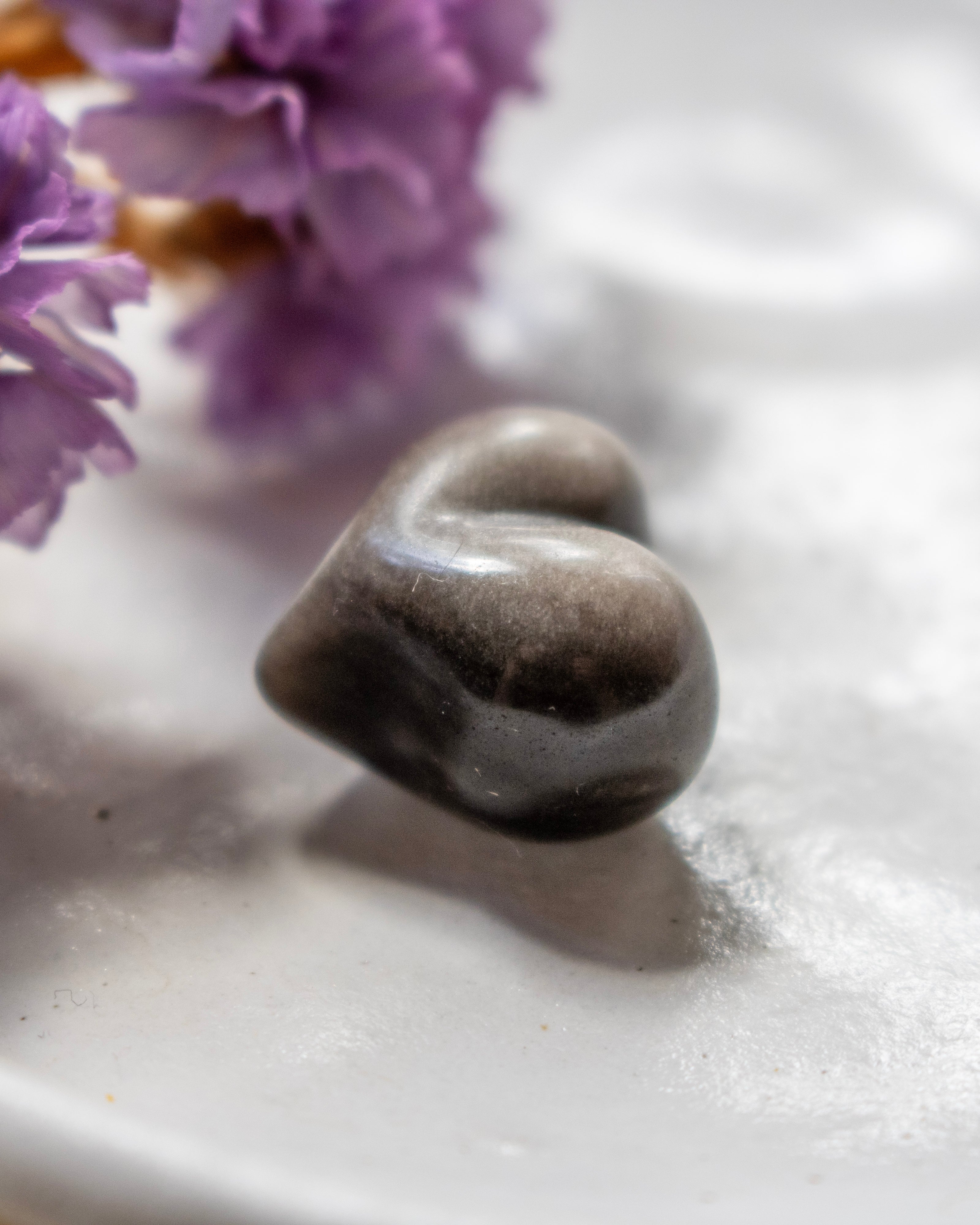 a black and silver heart shape stone on a white plate with soft shadows and purple flowers in the top left corner.