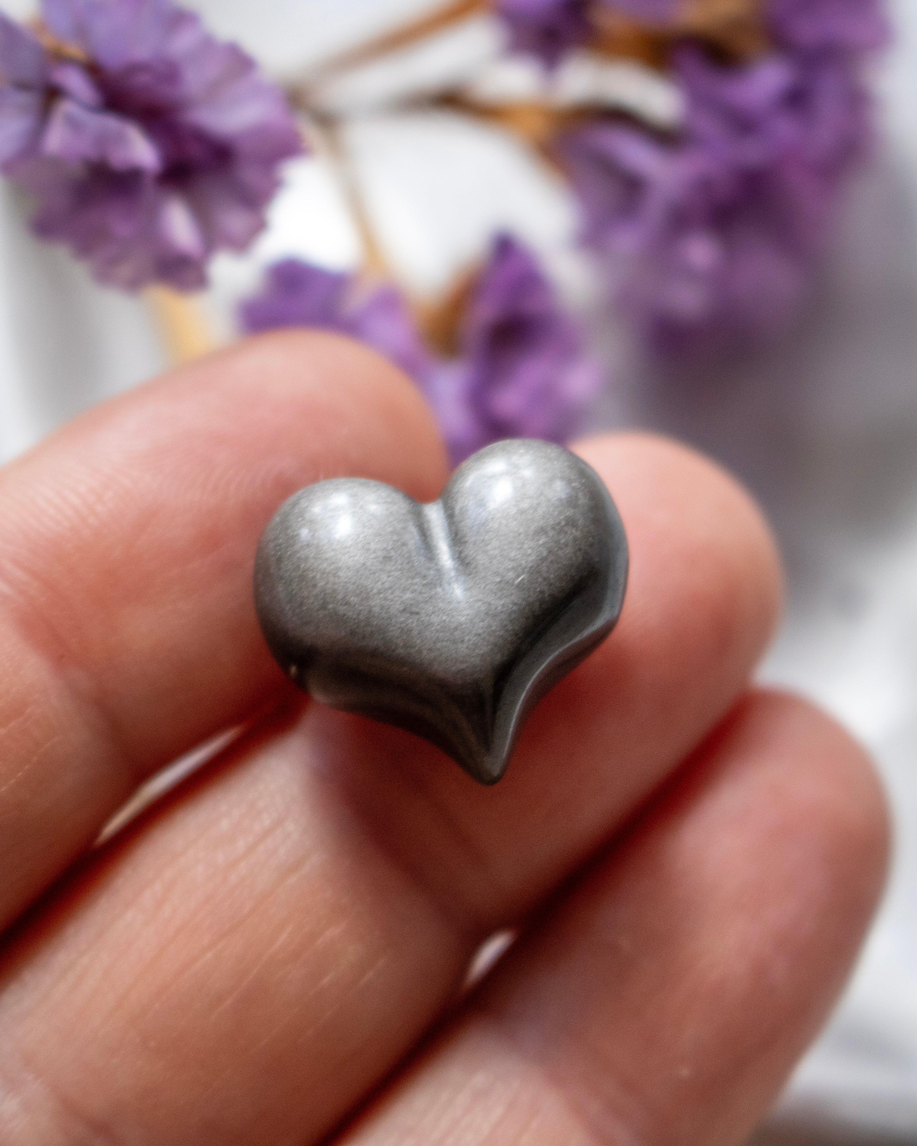 a black and silver heart shaped stone being held on finger tips with blurred purple flowers in the background