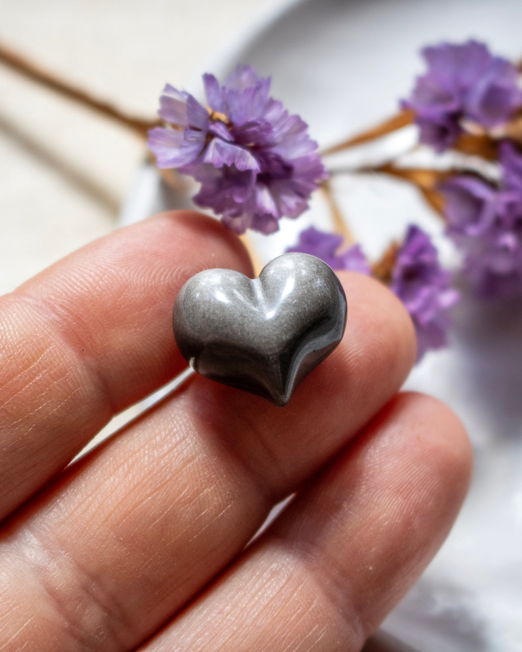 a black and silver heart shaped stone being held on finger tips with blurred purple flowers in the background