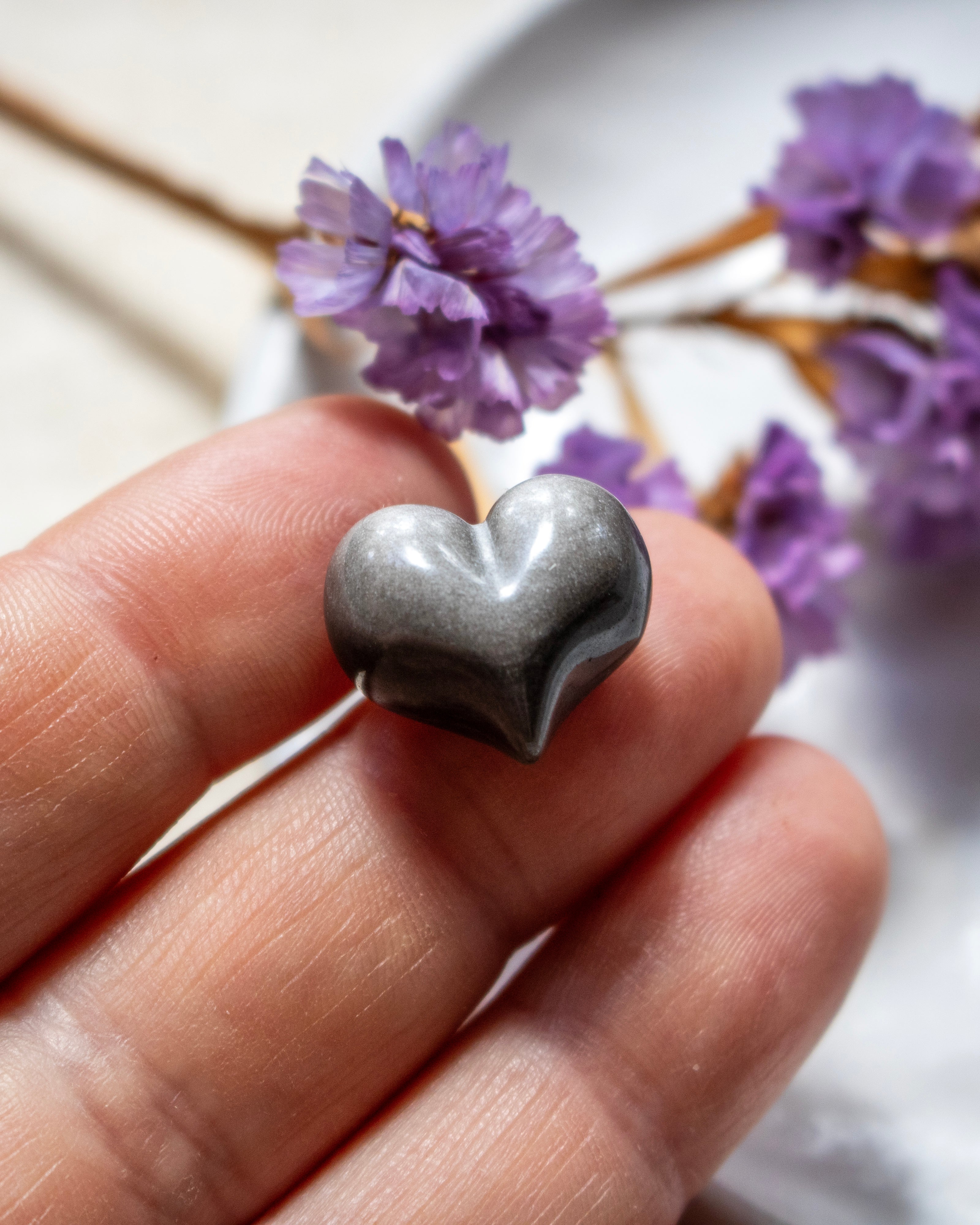 a black and silver heart shaped stone being held on finger tips with blurred purple flowers in the background