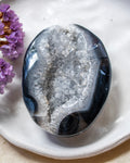 A polished geode stone with a dark blue-black outer shell and a white crystalline interior, displayed on a white ceramic dish with small purple flowers at the side.
