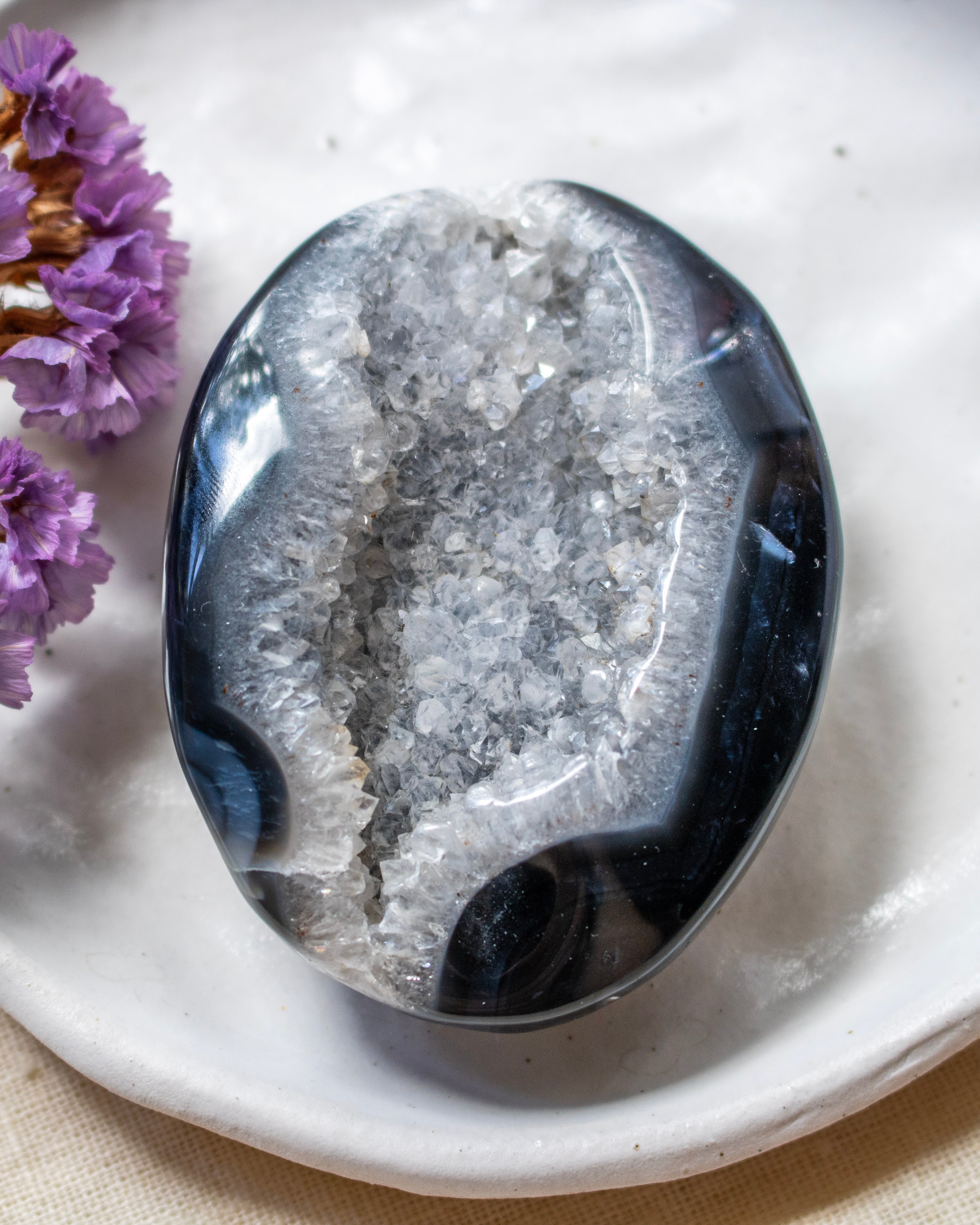 A polished geode stone with a dark blue-black outer shell and a white crystalline interior, displayed on a white ceramic dish with small purple flowers at the side.