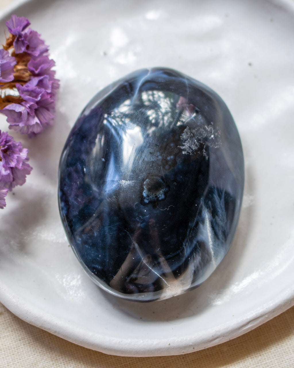 A polished dark blue and black stone with white marbled veins, resting on a white ceramic plate with dried purple flowers on the side.