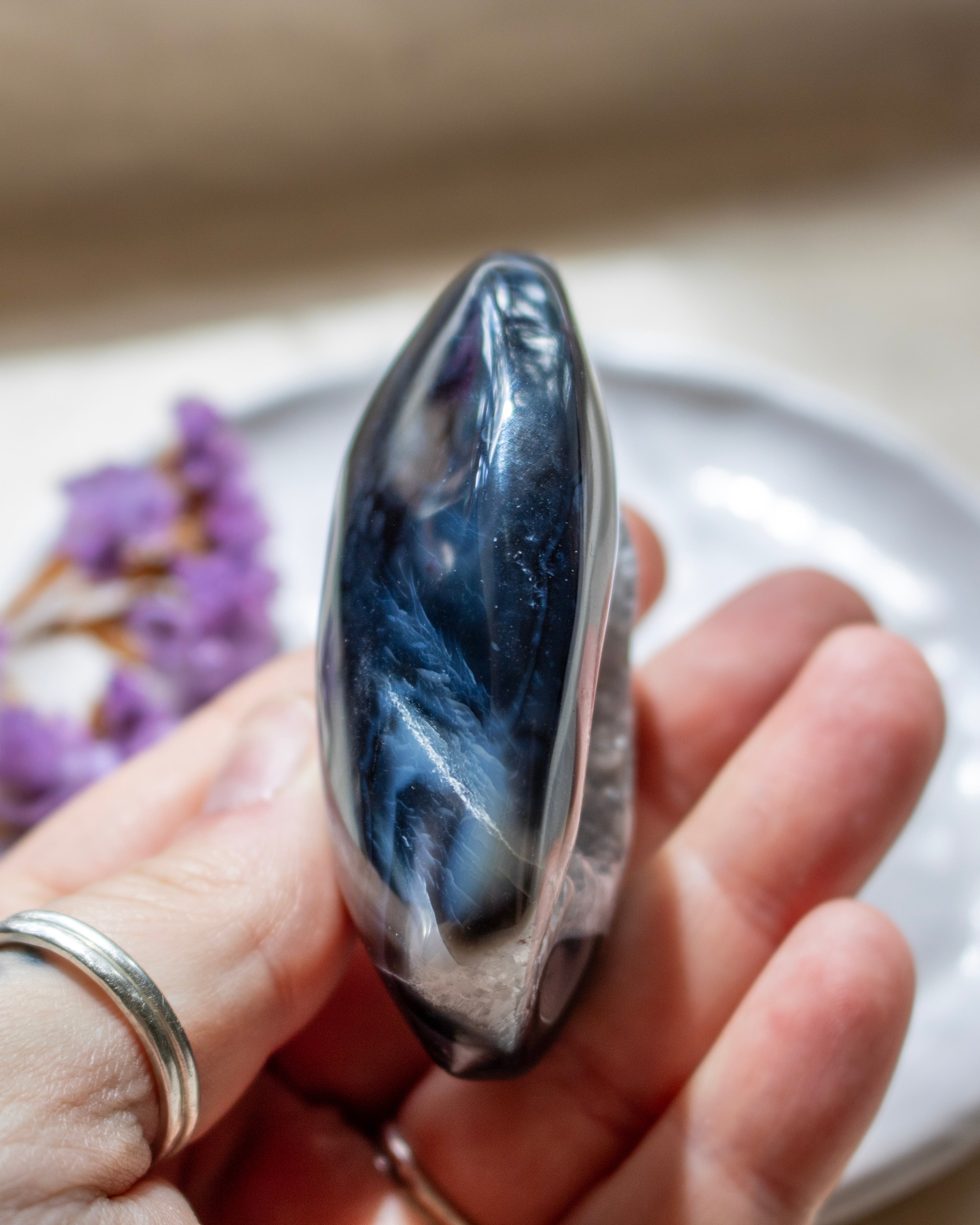 A polished dark blue and black stone held in a person's hand, with a soft-focus background showing a white plate and small purple flowers.