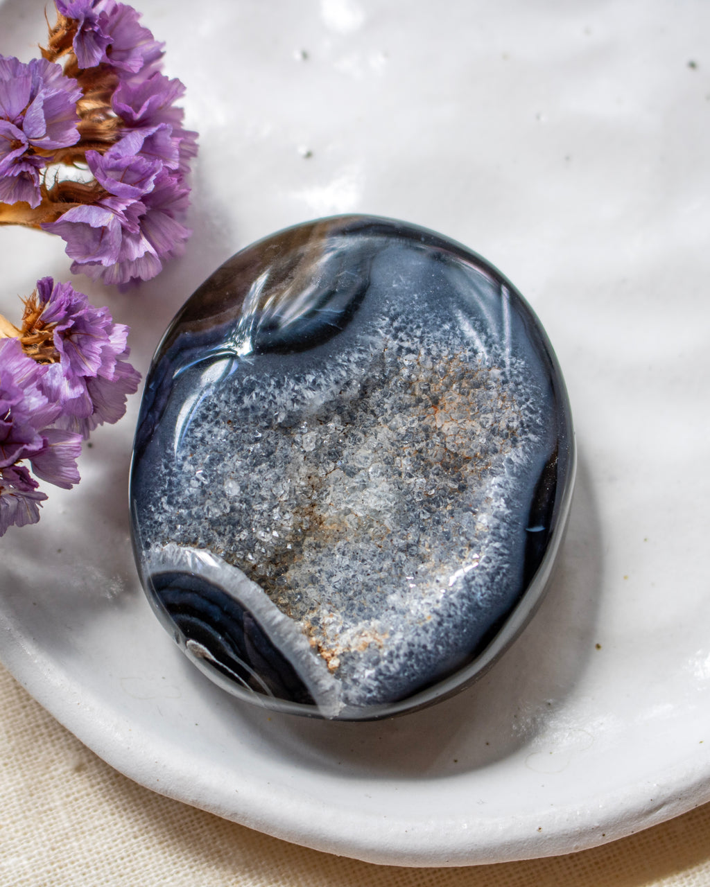 A polished dark blue and gray geode stone with white and gold crystalline formations, resting on a white ceramic dish with dried purple flowers in the corner.