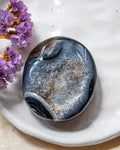 A polished dark blue and gray geode stone with white and gold crystalline formations, resting on a white ceramic dish with dried purple flowers in the corner.