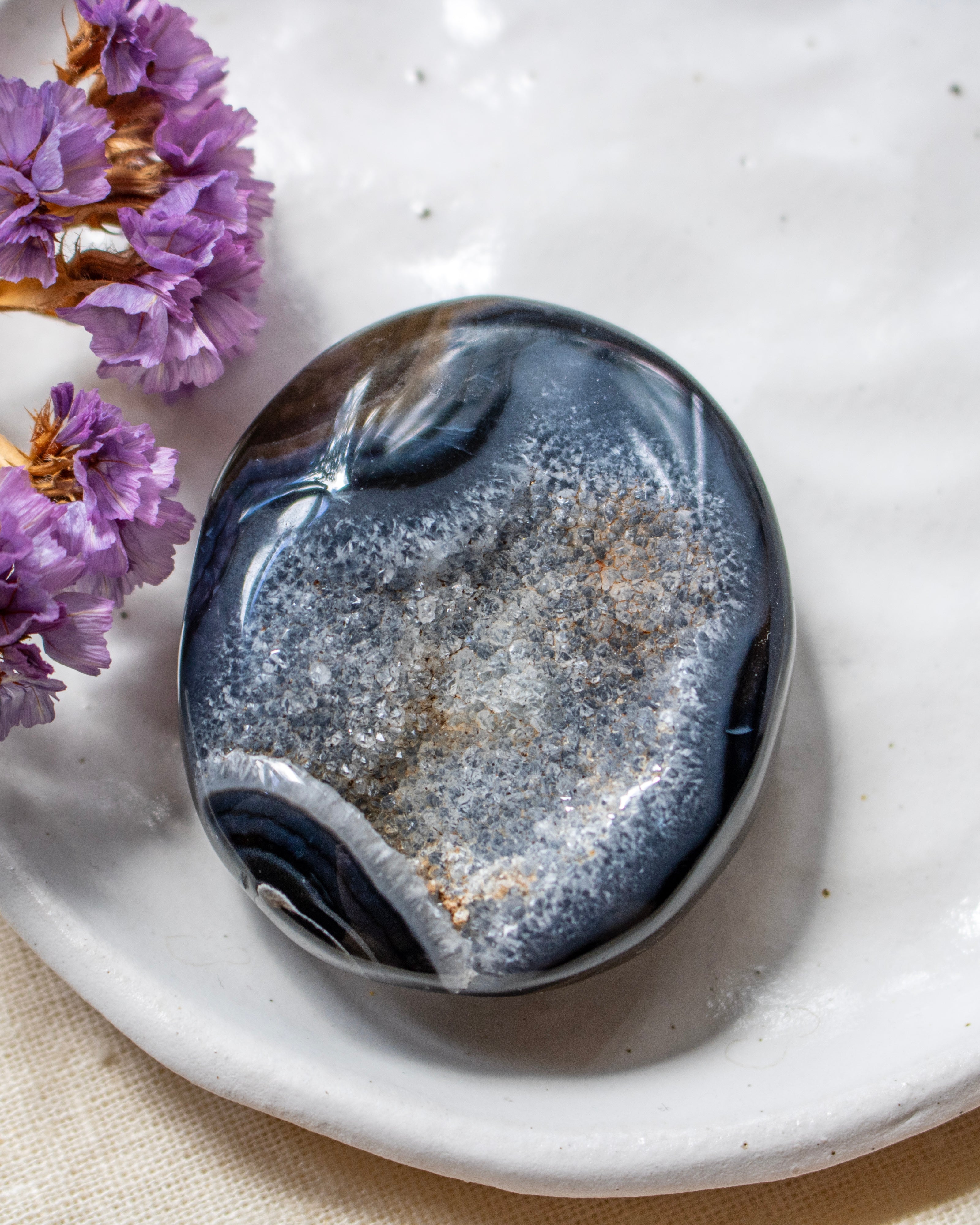A polished dark blue and gray geode stone with white and gold crystalline formations, resting on a white ceramic dish with dried purple flowers in the corner.