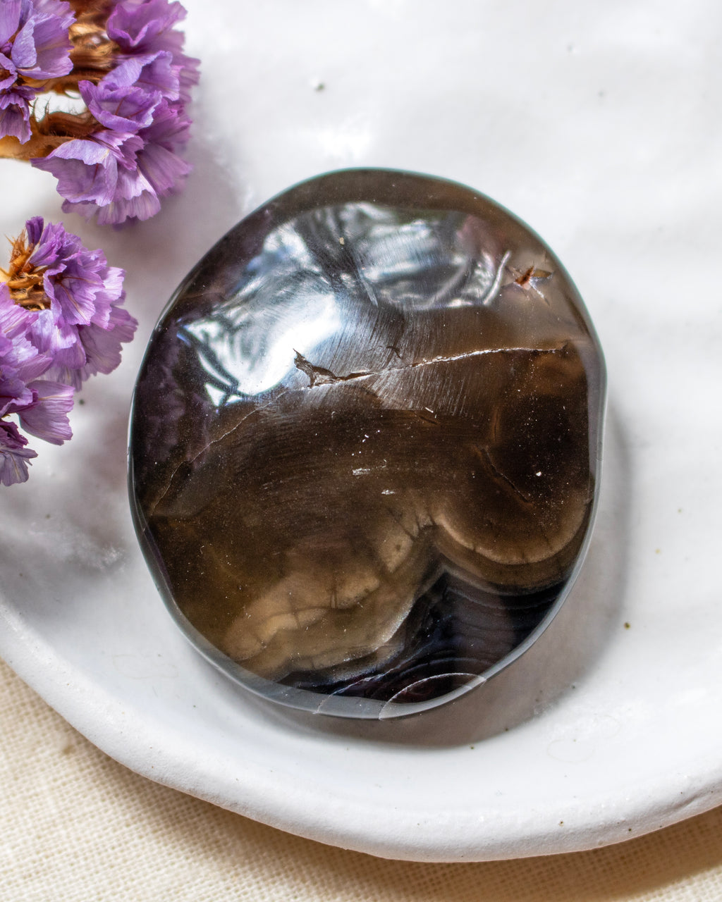 A polished dark brown smoky stone cabochon resting on a white ceramic dish, with dried purple flowers in the corner.