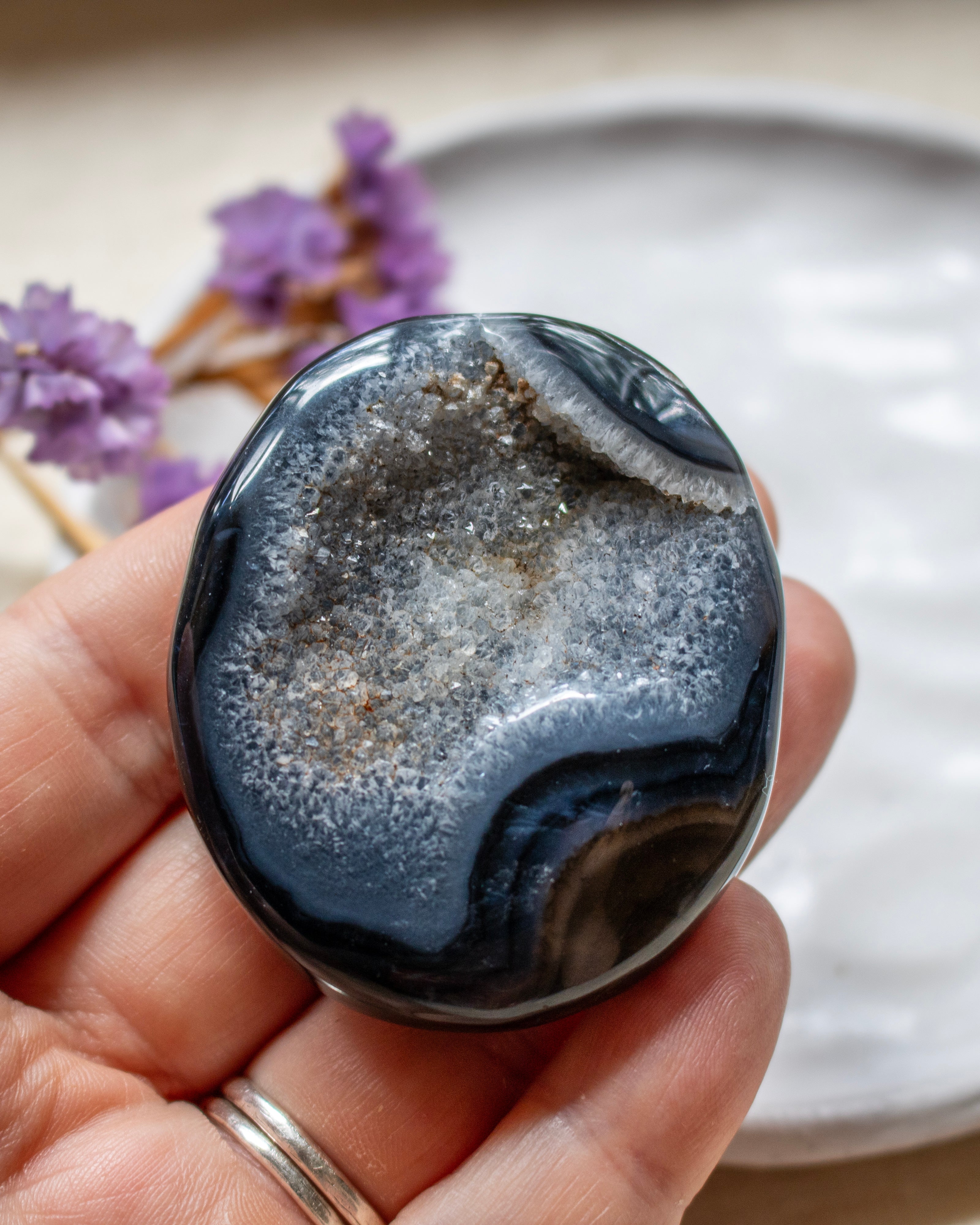 A polished round agate stone with a dark blue-gray outer band and a white crystalline center, held in a person's hand. The background is softly blurred with a white plate and small purple flowers.