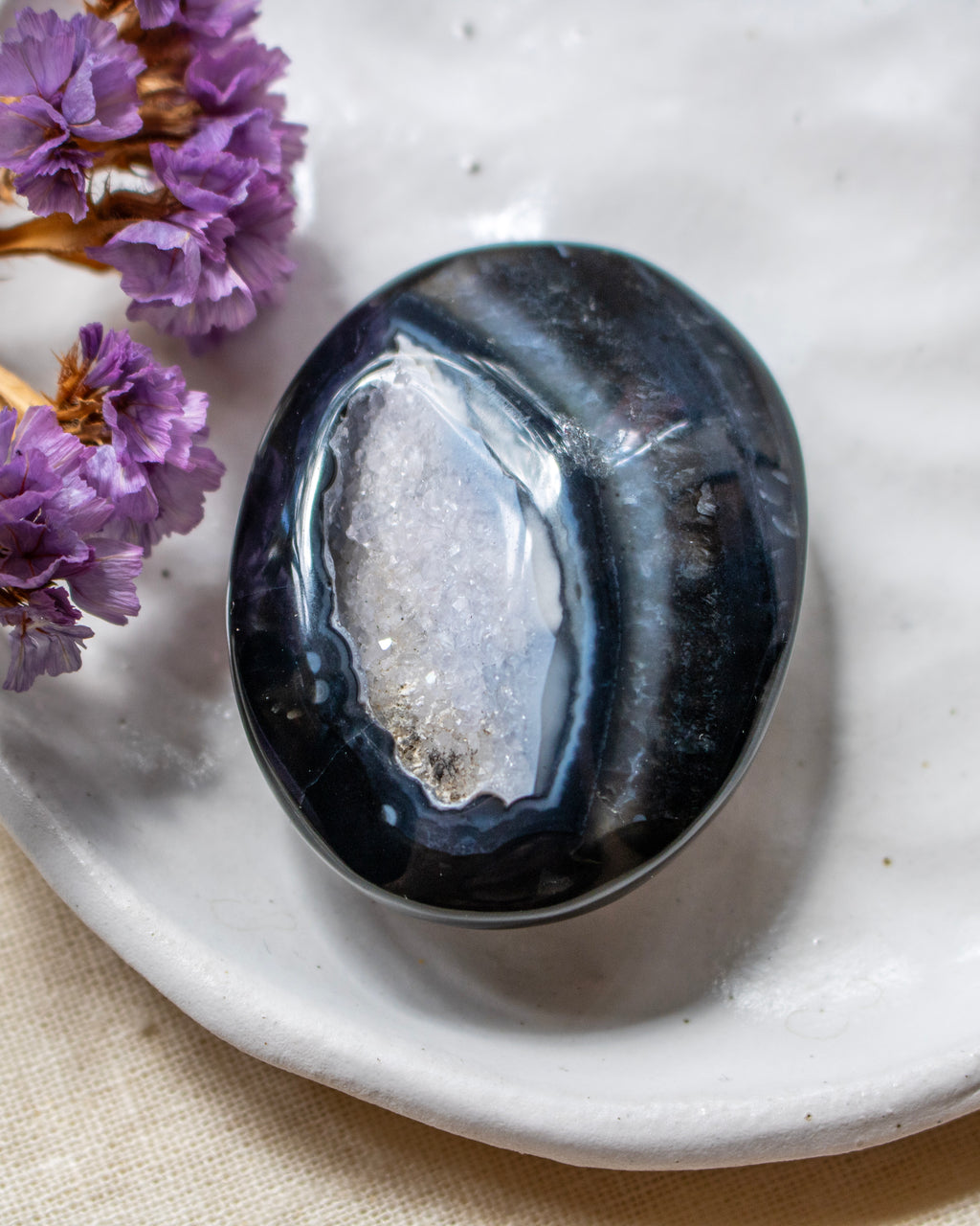 A polished black agate crystal stone with a white geode druzy interior, resting on a textured white ceramic dish with dried purple flowers in the corner.