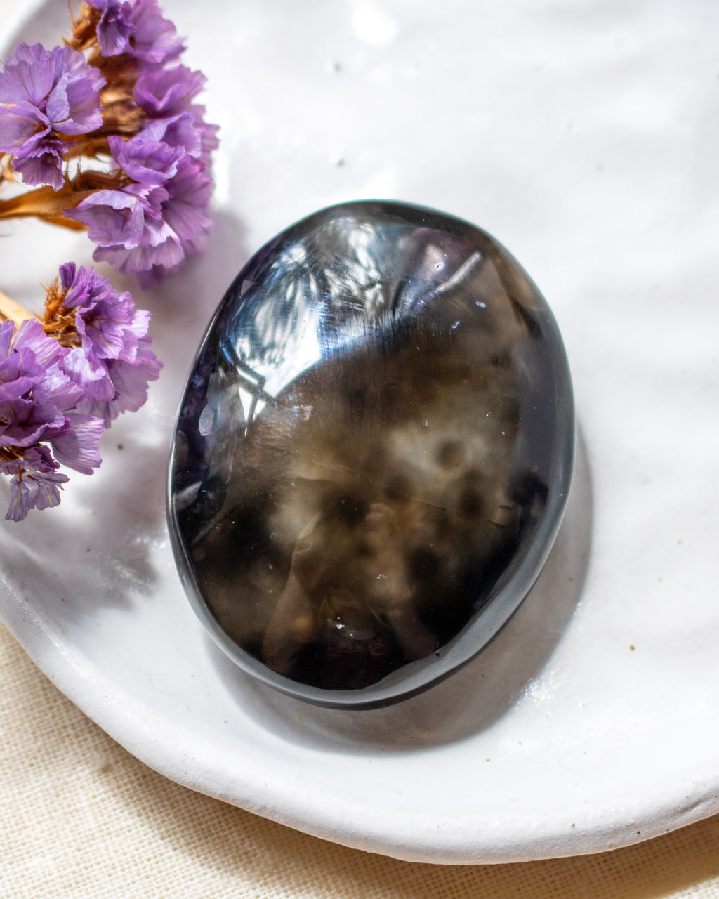 A polished dark smoky stone cabochon with natural inclusions, resting on a white ceramic dish next to dried purple flowers.