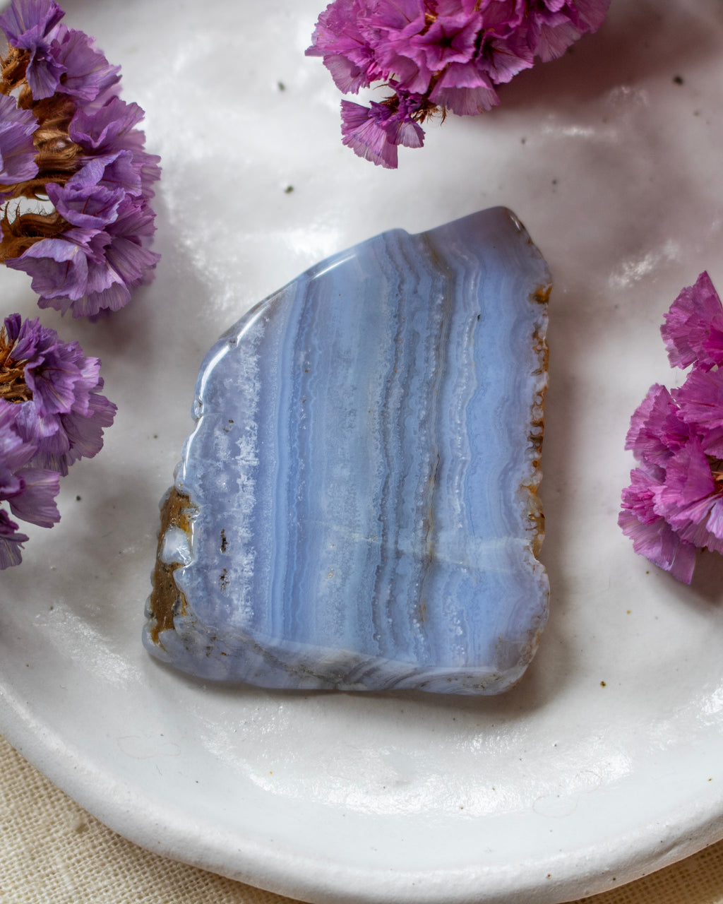 A polished blue agate stone with natural banding and rough edges, displayed on a white ceramic plate surrounded by small purple flowers.