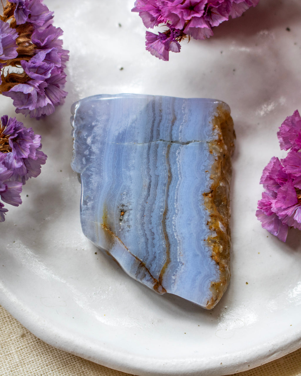 A polished natural stone slab with layered light blue, white, and brown veining, displayed on a white ceramic plate surrounded by small purple flowers.