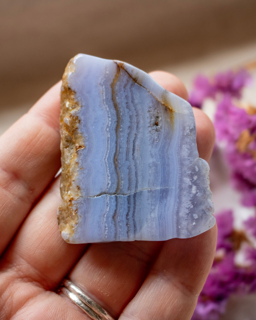 A hand holding a polished natural agate crystal with layered light blue bands, brown matrix, and a white crystal cluster on one end, with blurred purple flowers in the background.