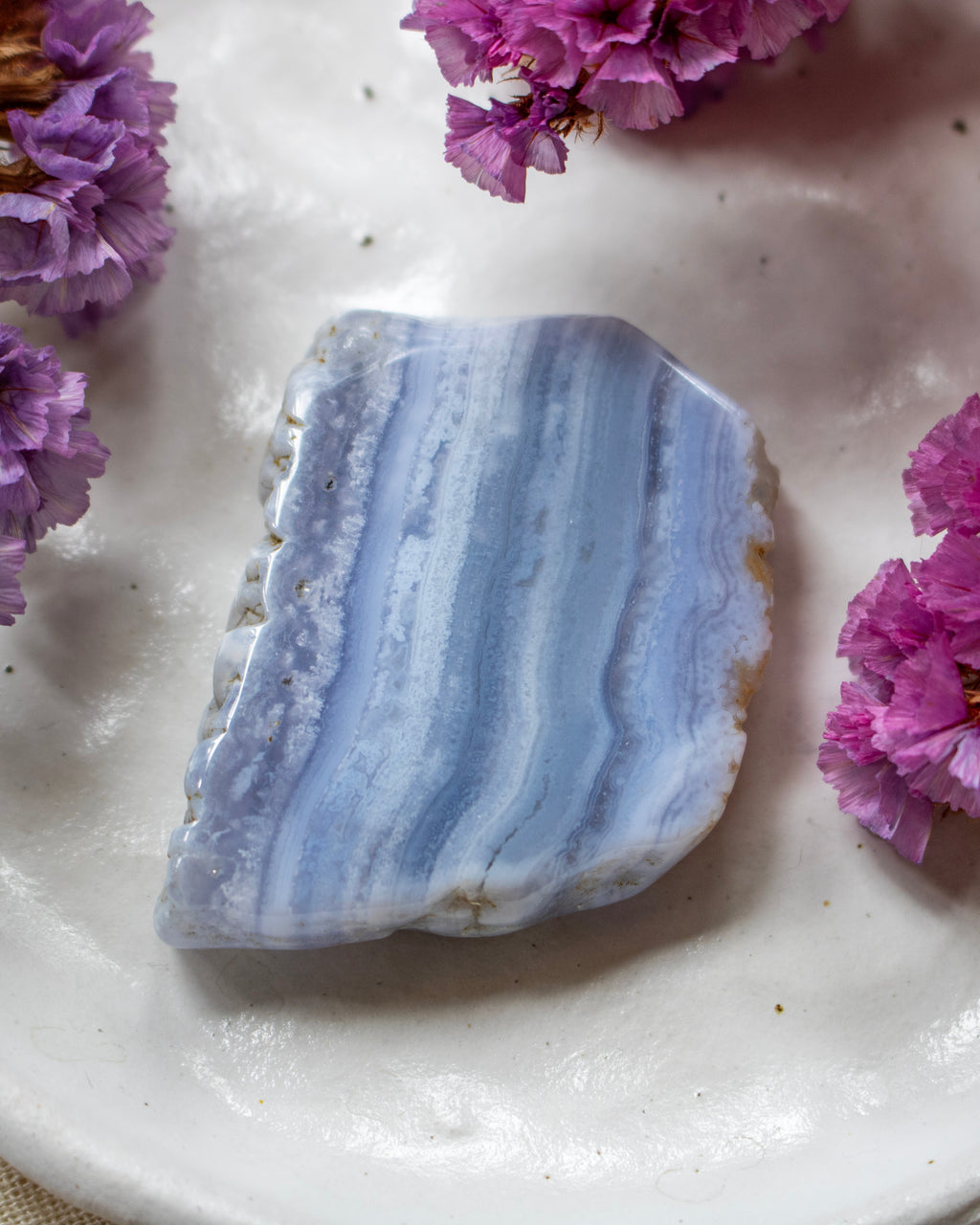 A polished natural stone slice with vertical light blue and white banding, displayed on a white ceramic plate with purple flowers around it.