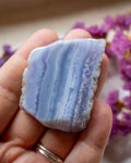 A hand holding a polished blue and white banded stone, likely a agate or marble slab, with purple flowers blurred in the background.