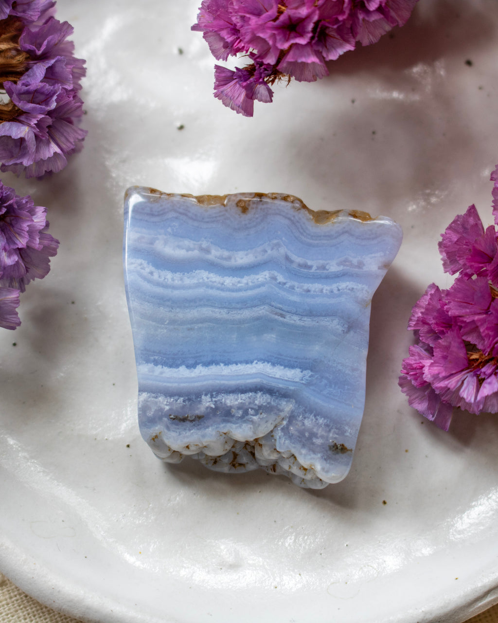 A polished blue and white banded stone slab, resembling agate or jasper, displayed on a white ceramic plate with purple flowers around it.