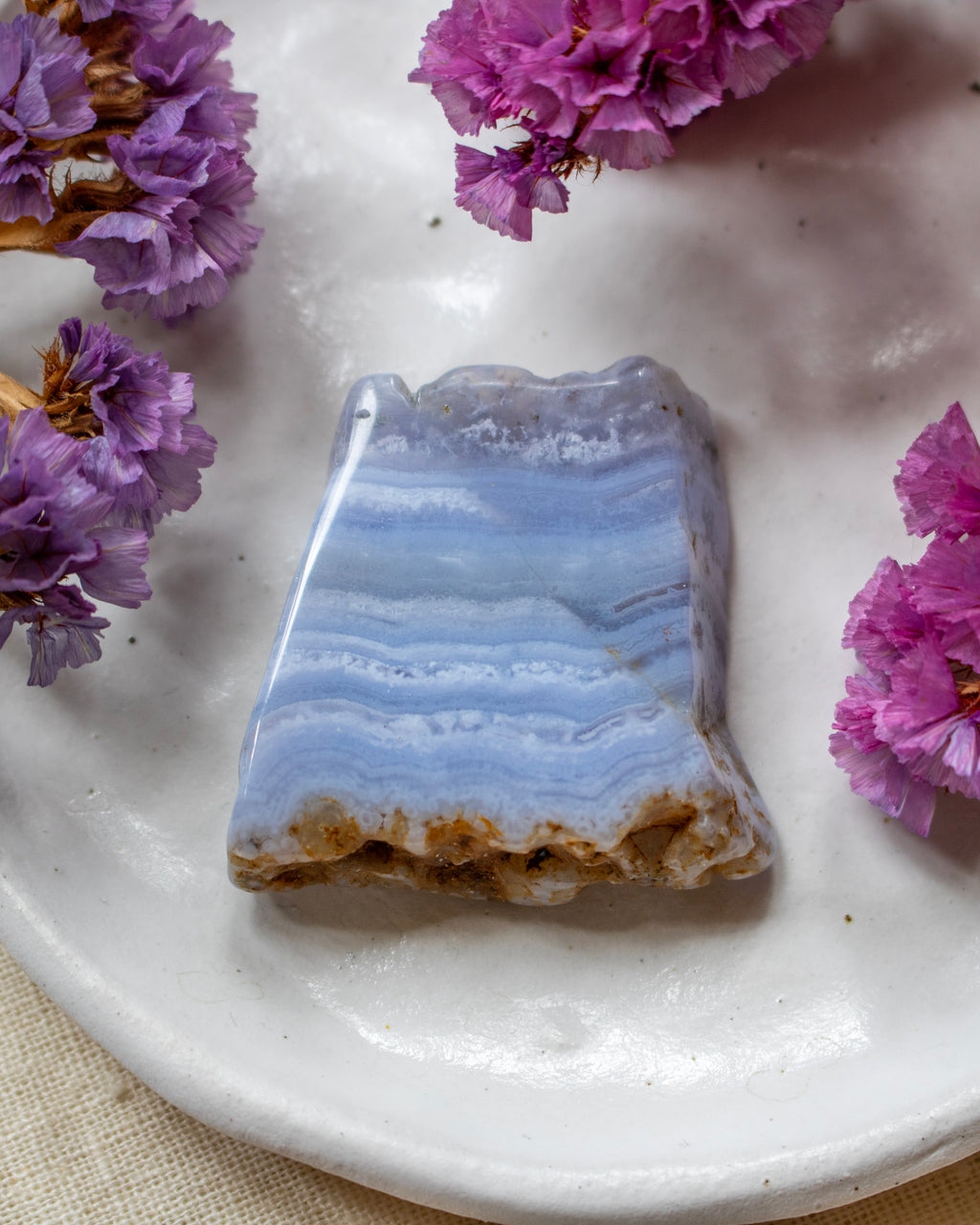 A polished irregular stone with layered light blue and white bands and a rough brown base, displayed on a white plate with purple flowers around it.