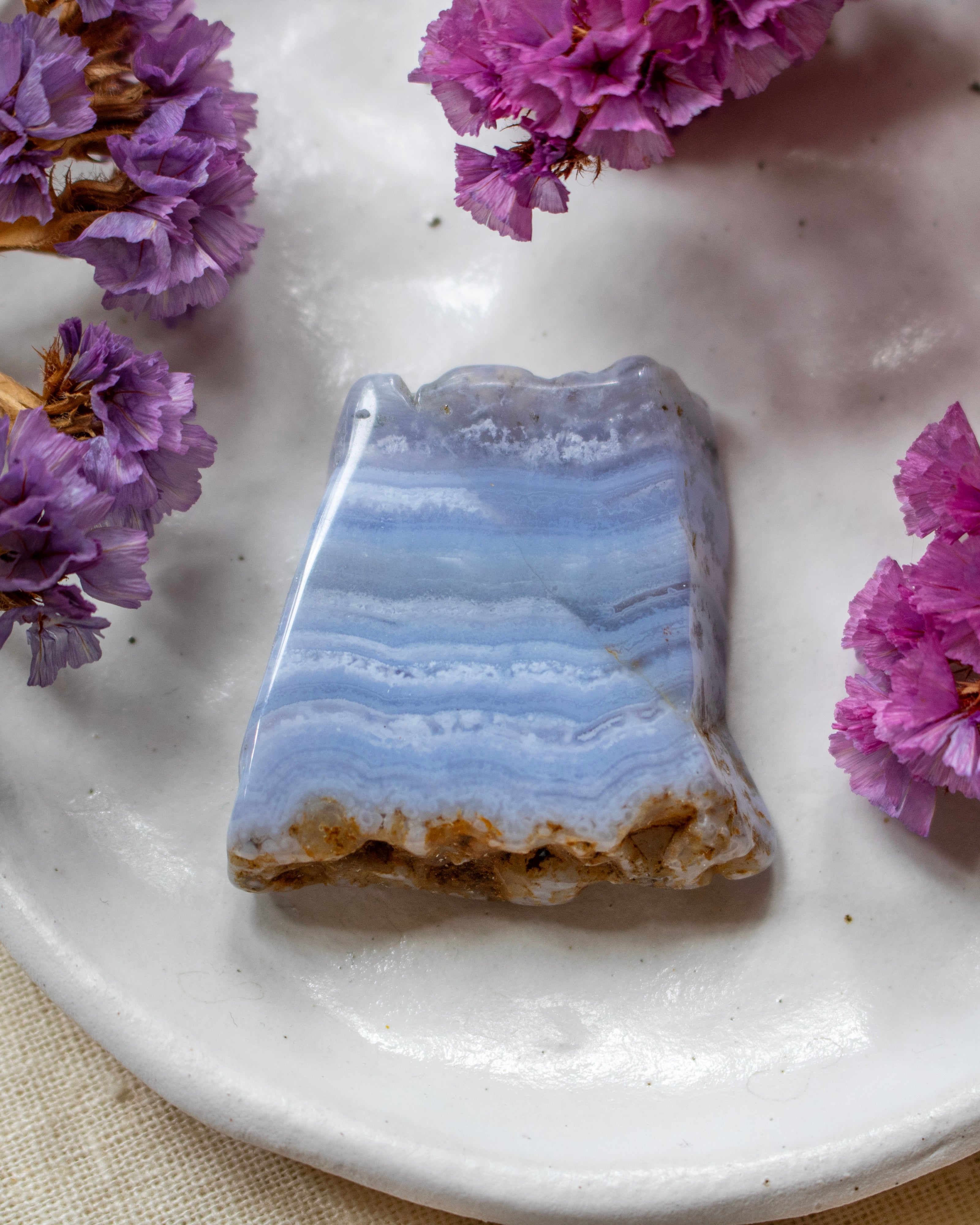 A polished irregular stone with layered light blue and white bands and a rough brown base, displayed on a white plate with purple flowers around it.