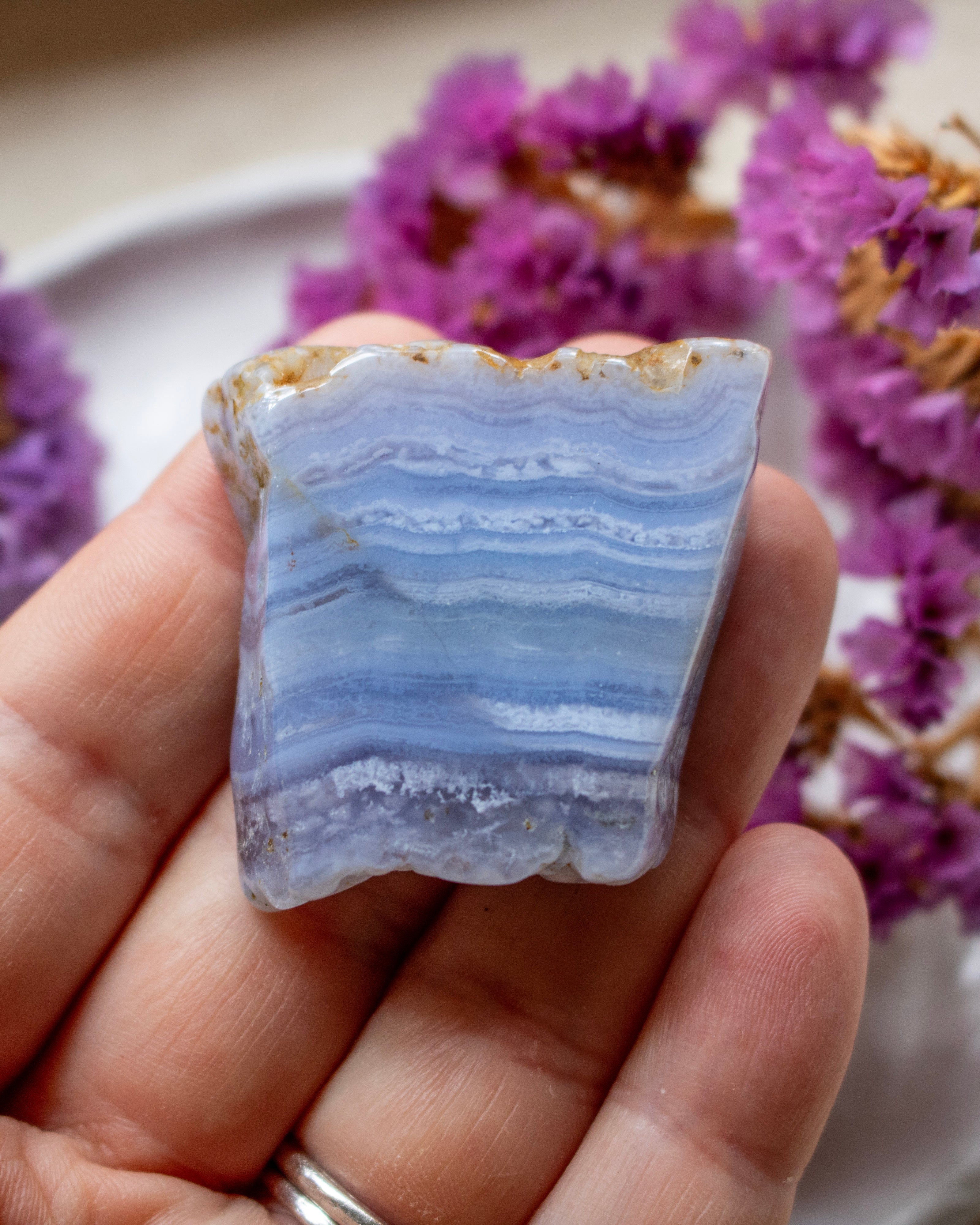 A hand holding a polished blue and white banded agate stone with natural rough edges, set against a soft-focus background of purple dried flowers.