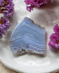 A polished irregular-shaped agate stone with layered light blue and white bands, displayed on a white ceramic plate surrounded by small purple flowers.