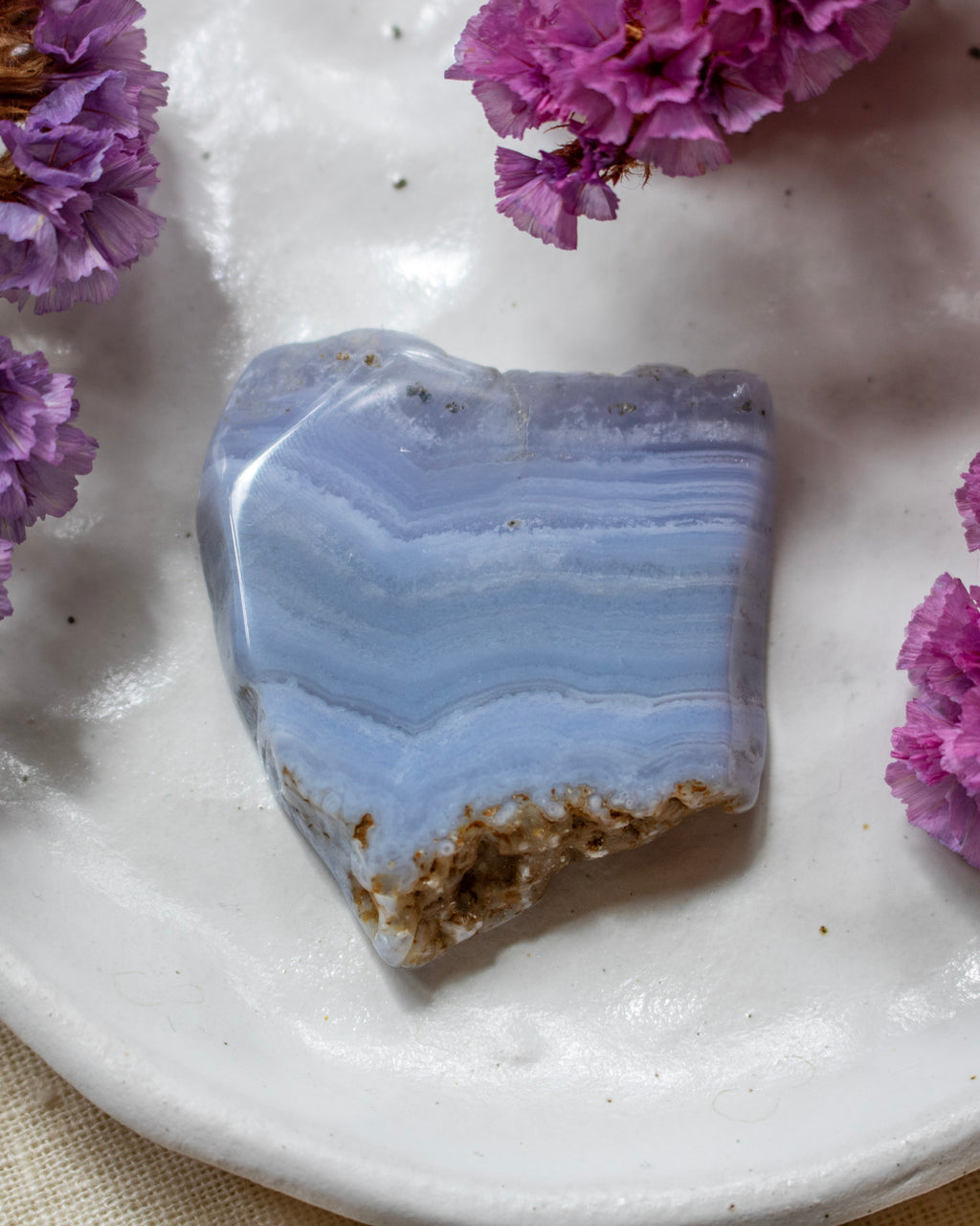 A polished piece of light blue and white banded agate stone with a rough, textured brown crystalline base, displayed on a white ceramic plate decorated with purple flowers.