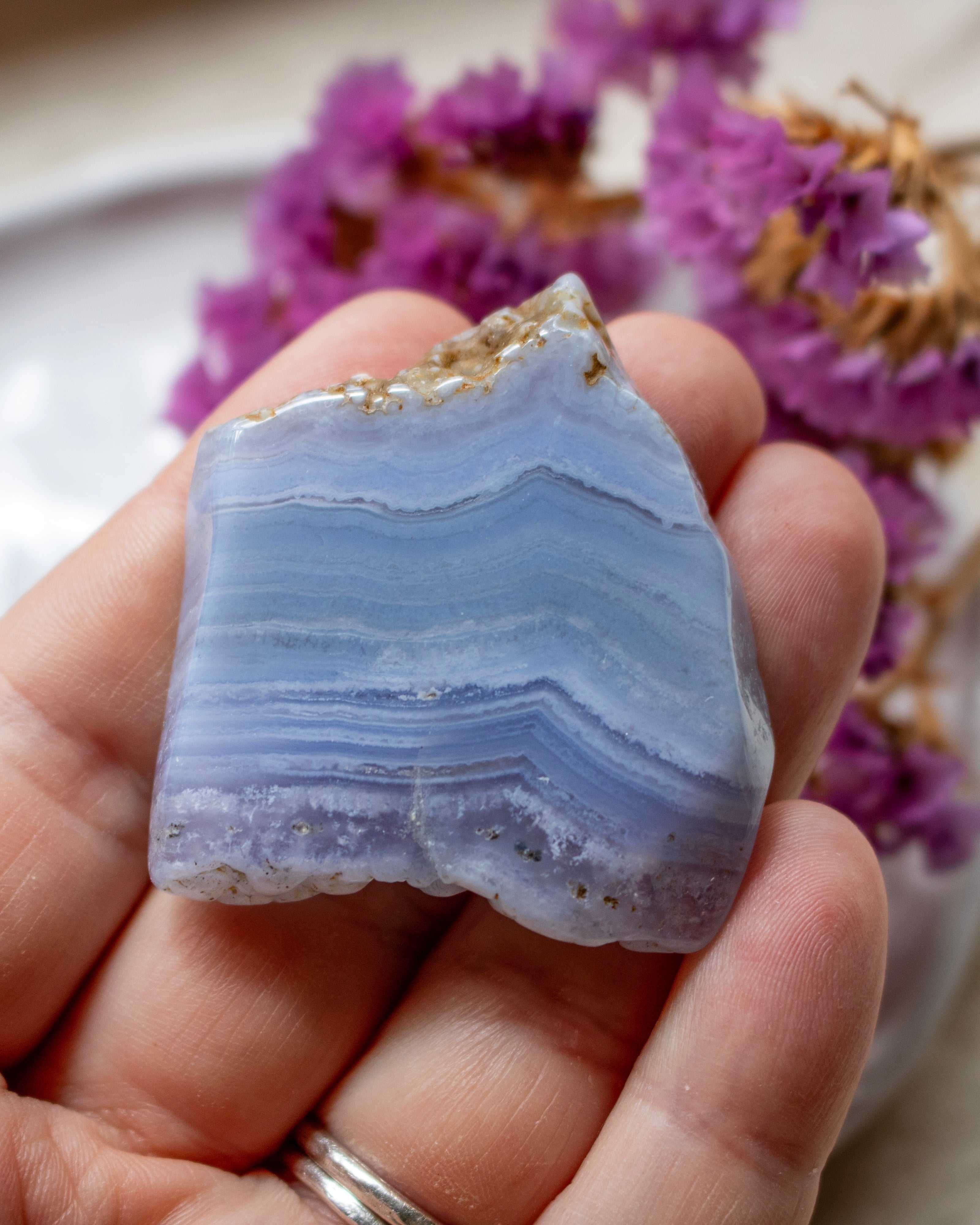 A hand holding a polished agate crystal specimen with layered light blue and lavender bands and a rough natural edge, with blurred purple flowers in the background.