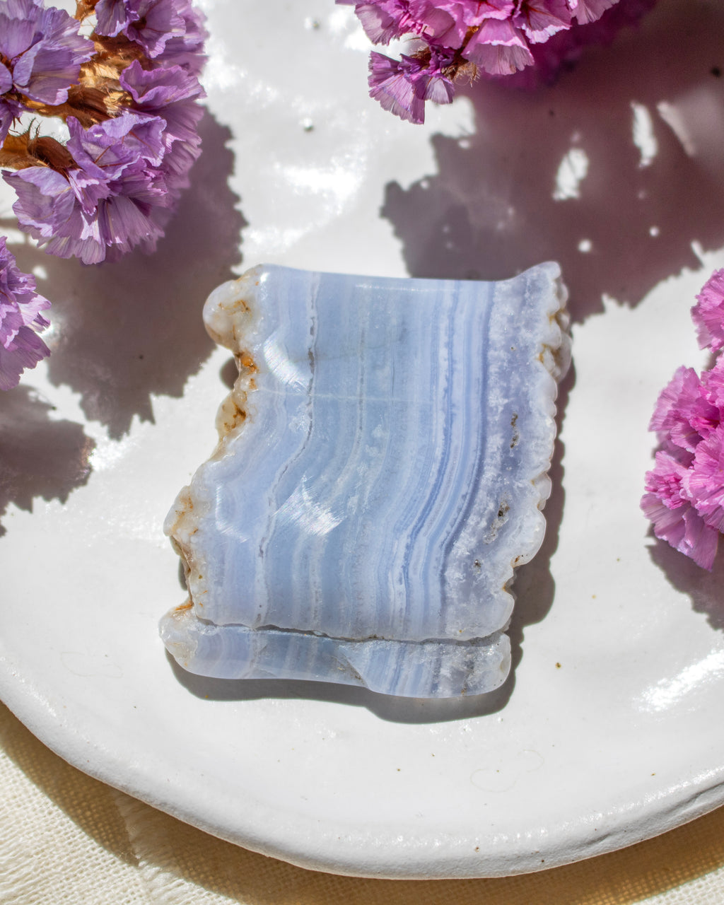 A polished blue agate crystal with natural rough edges, resting on a white ceramic plate decorated with dried pink flowers under bright sunlight.