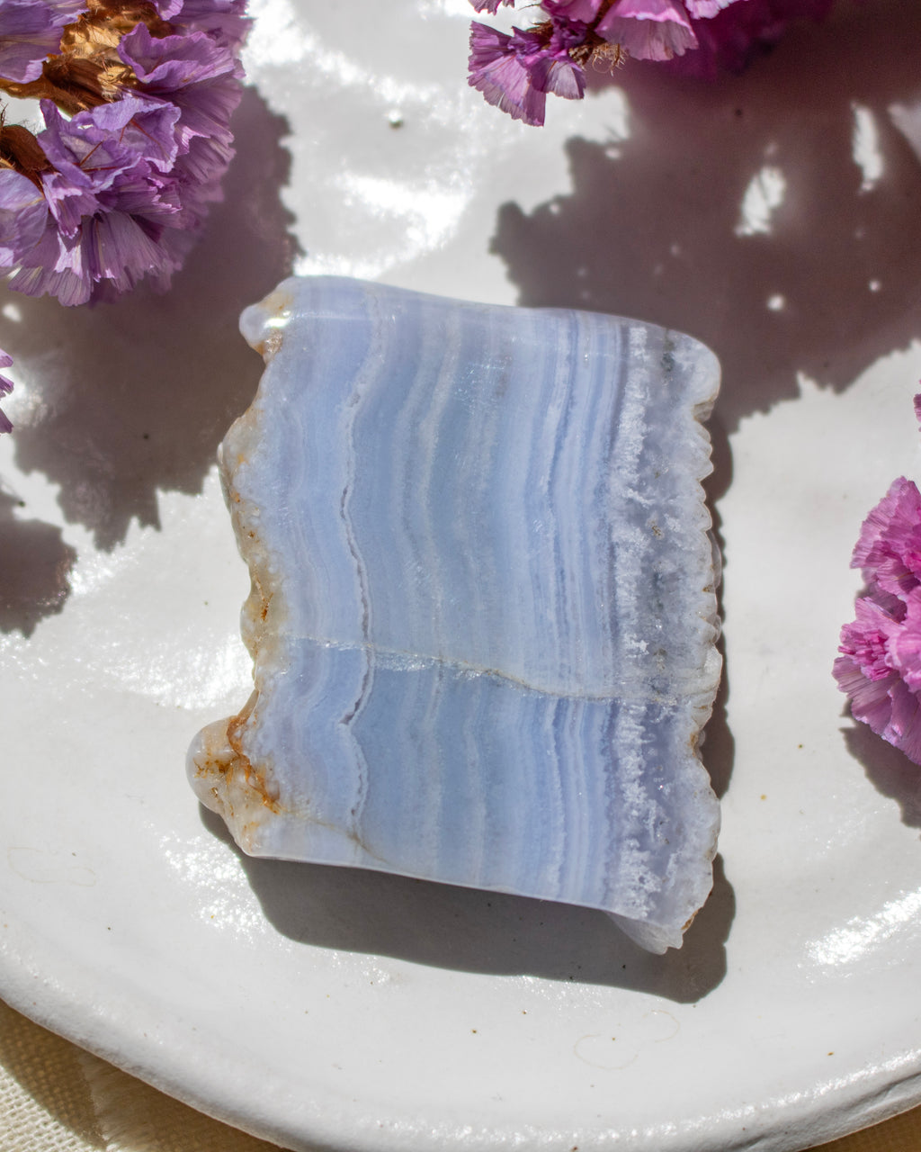 A polished rectangular stone slice with pale blue and white banded striations, placed on a white plate with purple flowers around it.