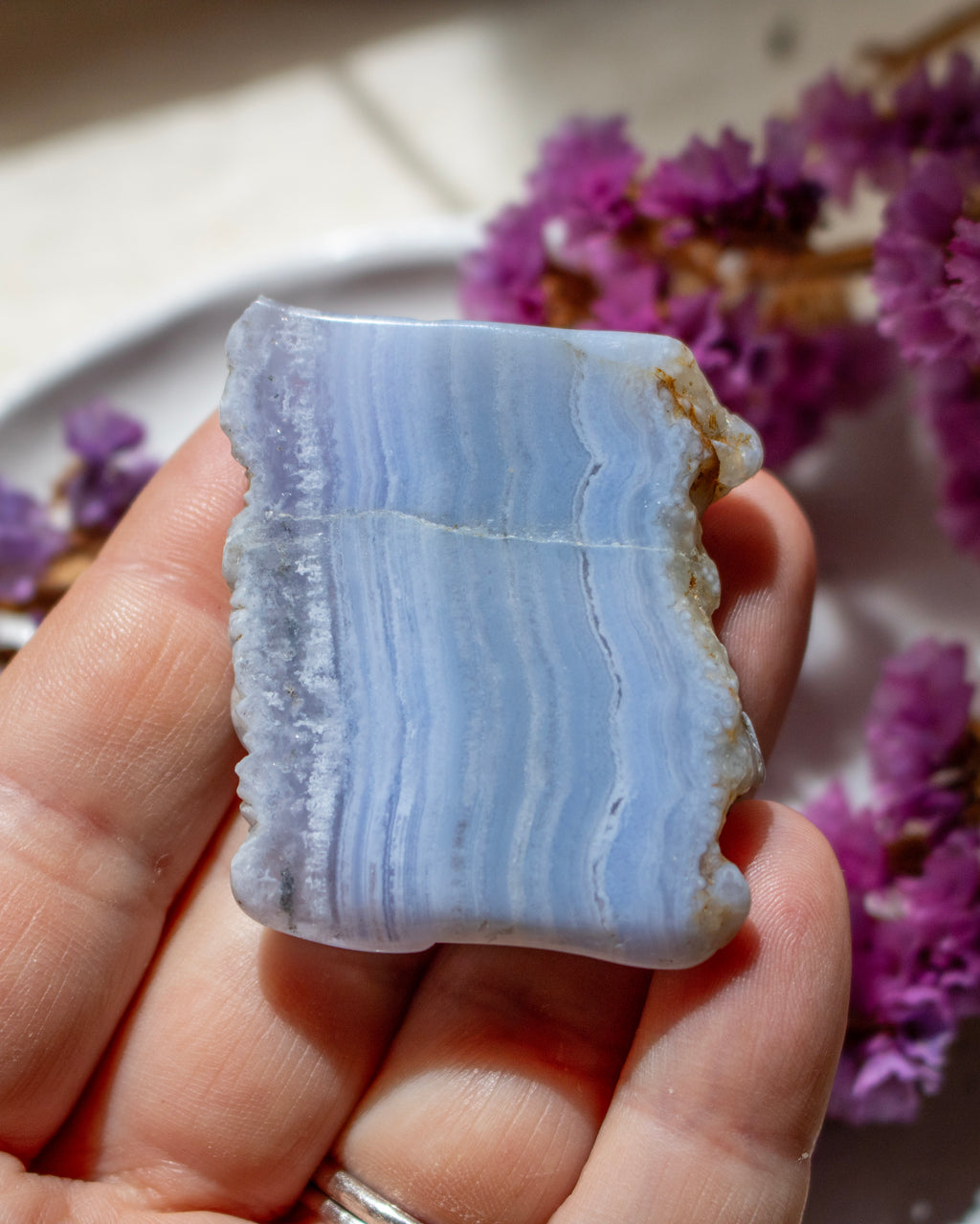 A hand holding a polished rectangular piece of pale blue and white banded stone, likely agate or marble, with rough natural edges, against a softly blurred background with purple flowers.