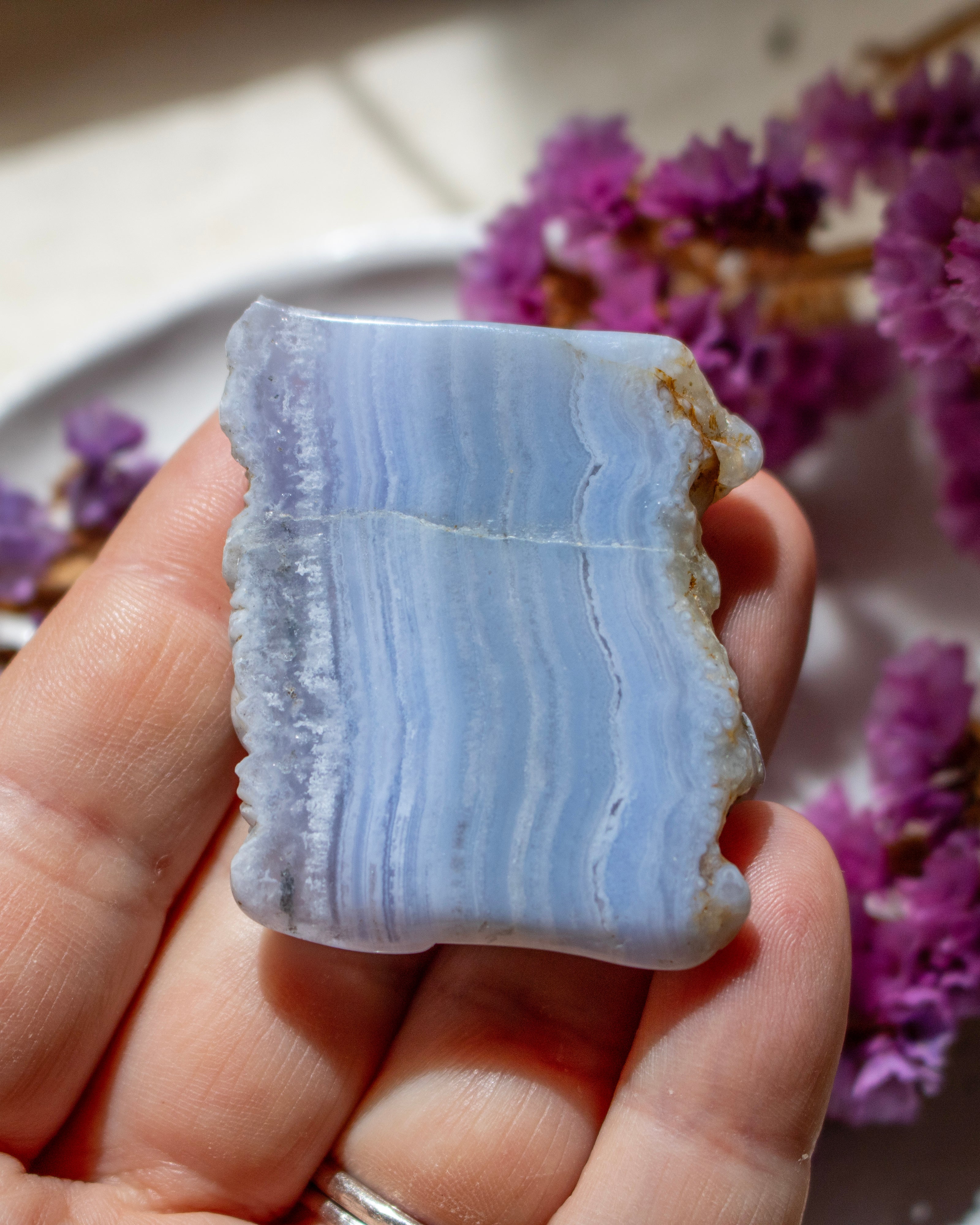 A hand holding a polished rectangular piece of pale blue and white banded stone, likely agate or marble, with rough natural edges, against a softly blurred background with purple flowers.