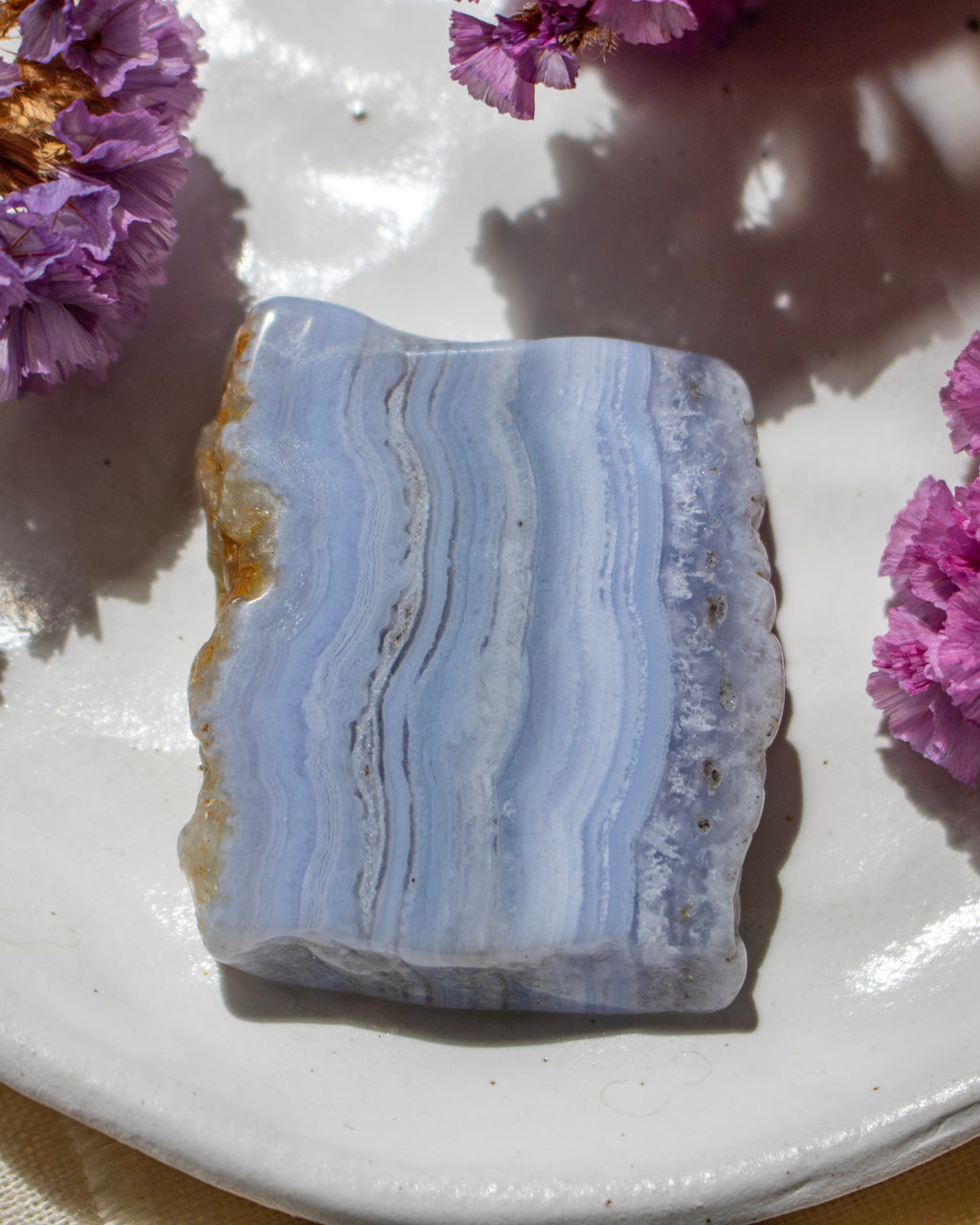 A polished rectangular stone slab with wavy light blue, white, and gray bands, placed on a white plate with purple flowers around it.