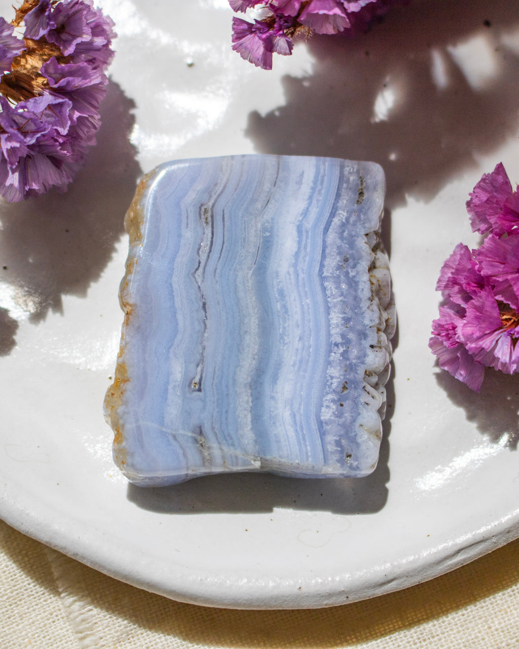 A polished rectangular stone slab with wavy light blue, white, and pale lavender banding, resting on a white ceramic plate with purple flowers around it.