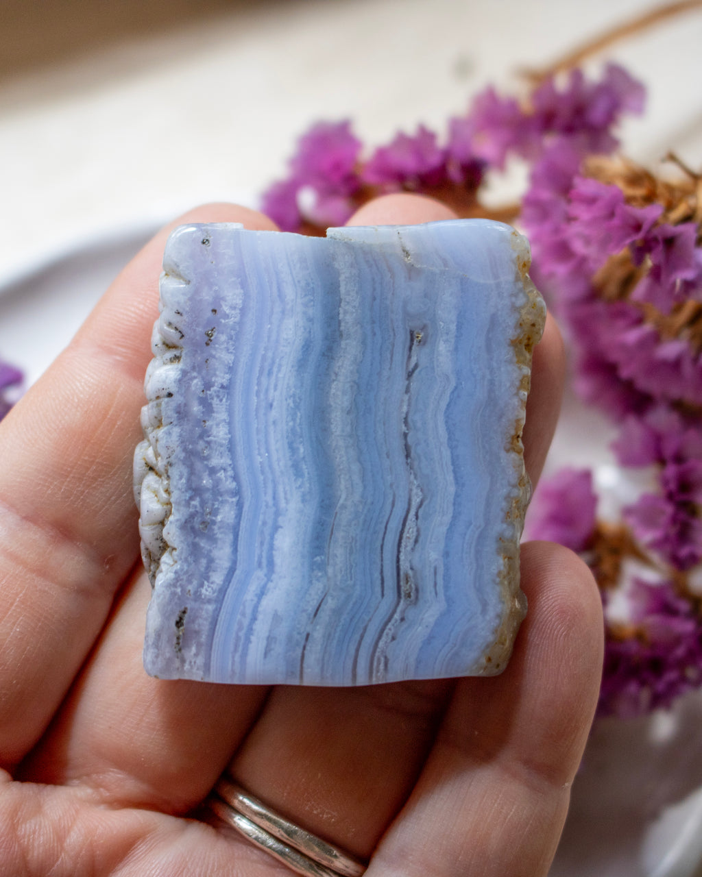 A hand holding a polished rectangular stone slice with pale blue and white banded stripes and rough natural edges, with dried purple flowers blurred in the background.