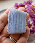 A hand holding a polished rectangular agate stone with horizontal lavender, blue, and gray banded stripes, with purple flowers blurred in the background.