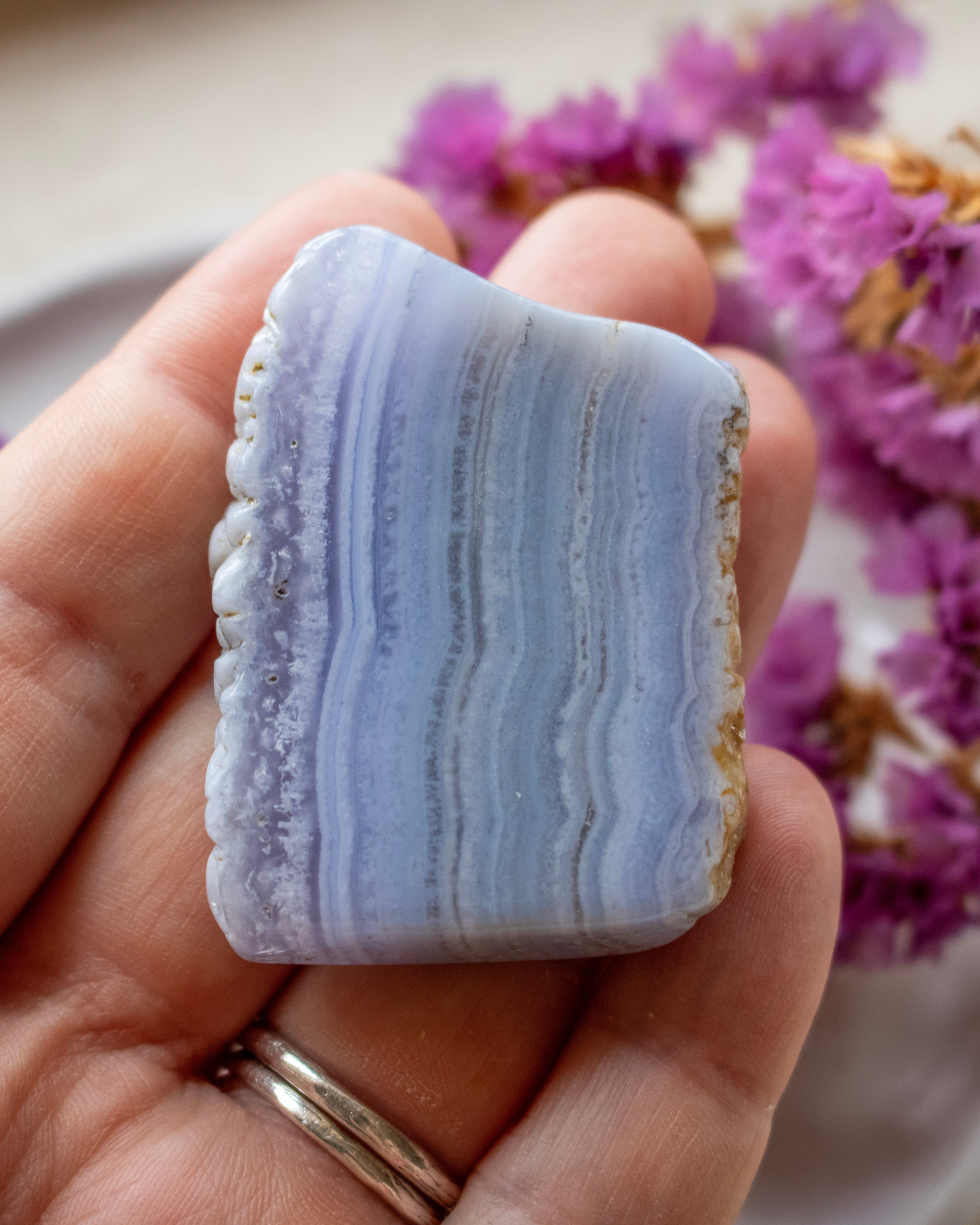 A hand holding a polished rectangular agate stone with horizontal lavender, blue, and gray banded stripes, with purple flowers blurred in the background.
