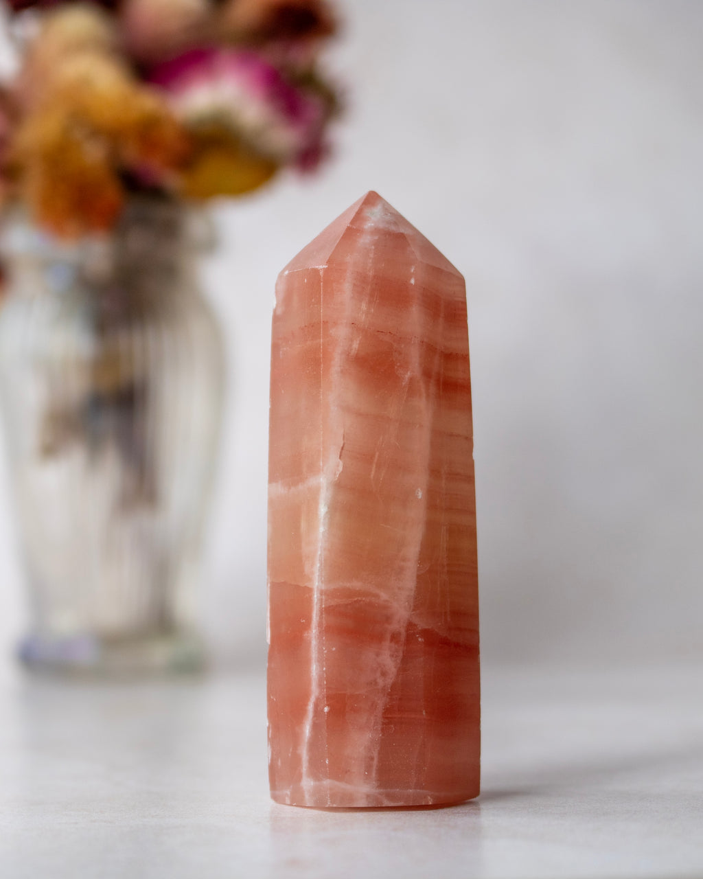 A natural rose quartz crystal tower with pointed tip stands on a light surface, with a blurred glass vase of dried flowers in the background.