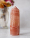 A tall, pointed pinkish-orange crystal tower with natural horizontal banding, standing on a light surface with a softly blurred floral vase in the background.