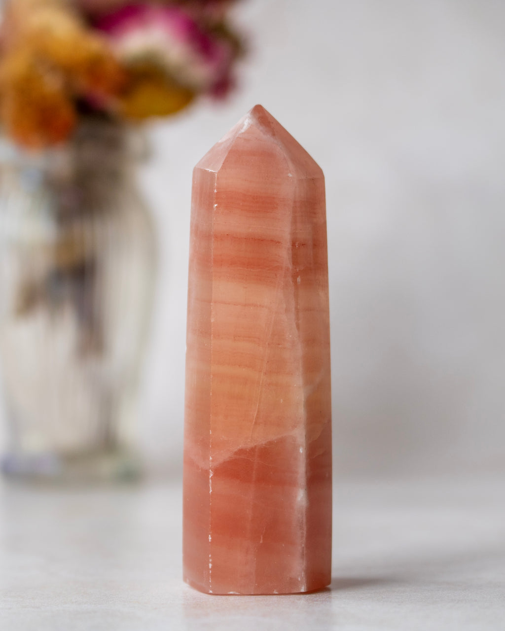 A natural rose quartz crystal tower with pointed top, standing upright on a light surface with a blurred floral vase in the background.