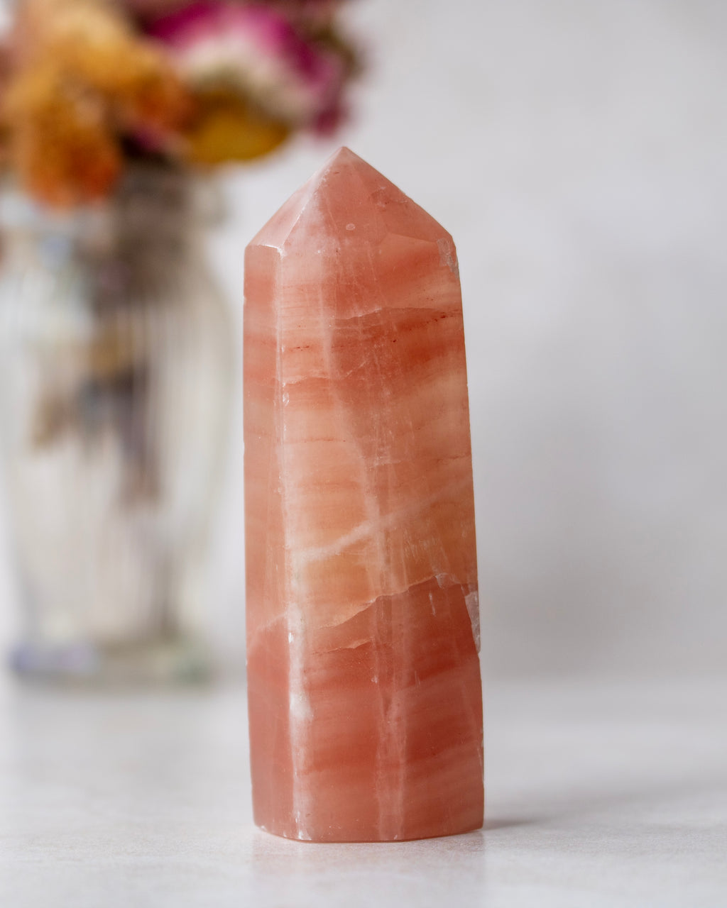 A polished peach-orange quartz point crystal tower standing upright on a light surface, with a softly blurred glass vase of dried flowers in the background.