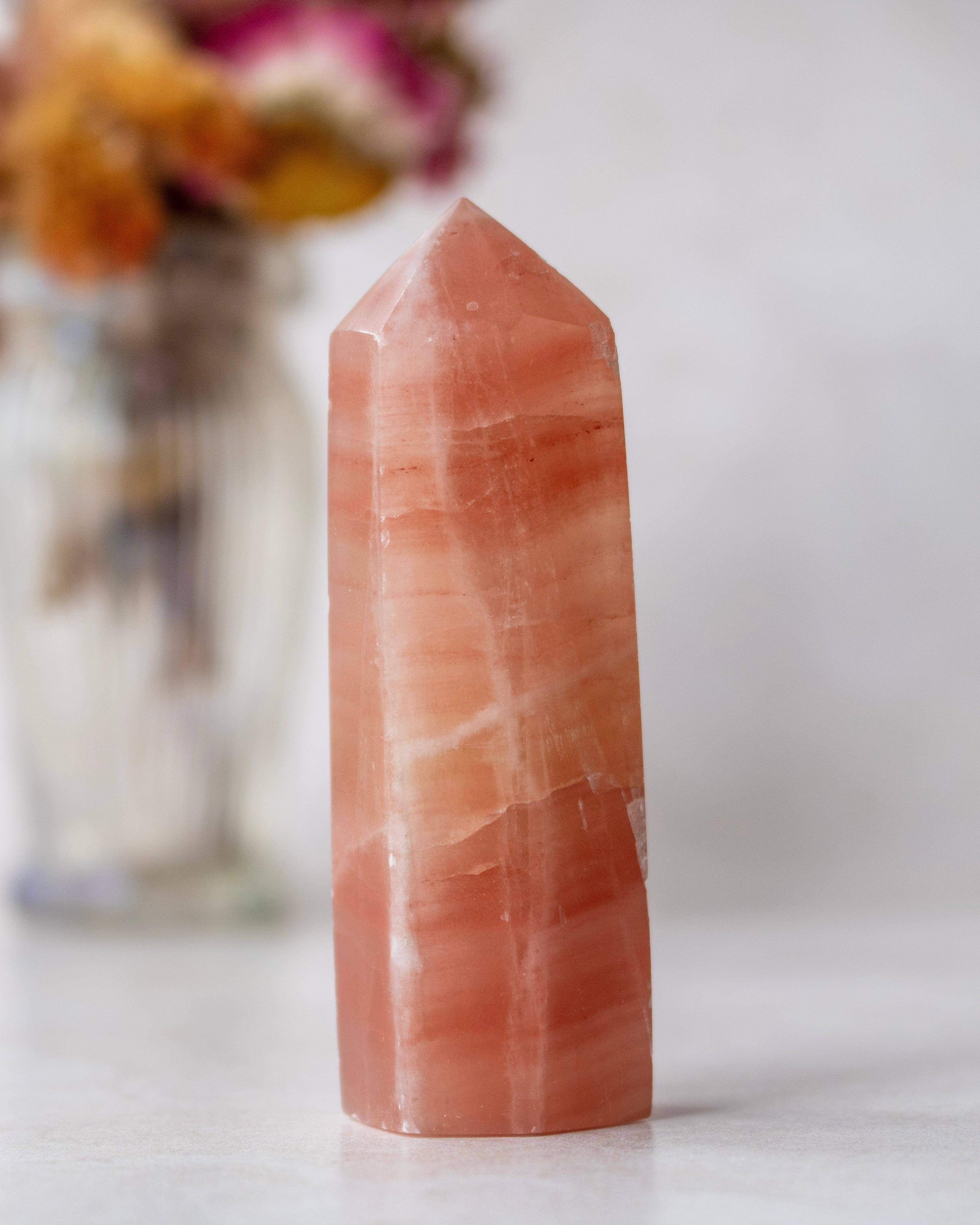 A polished peach-orange quartz point crystal tower standing upright on a light surface, with a softly blurred glass vase of dried flowers in the background.