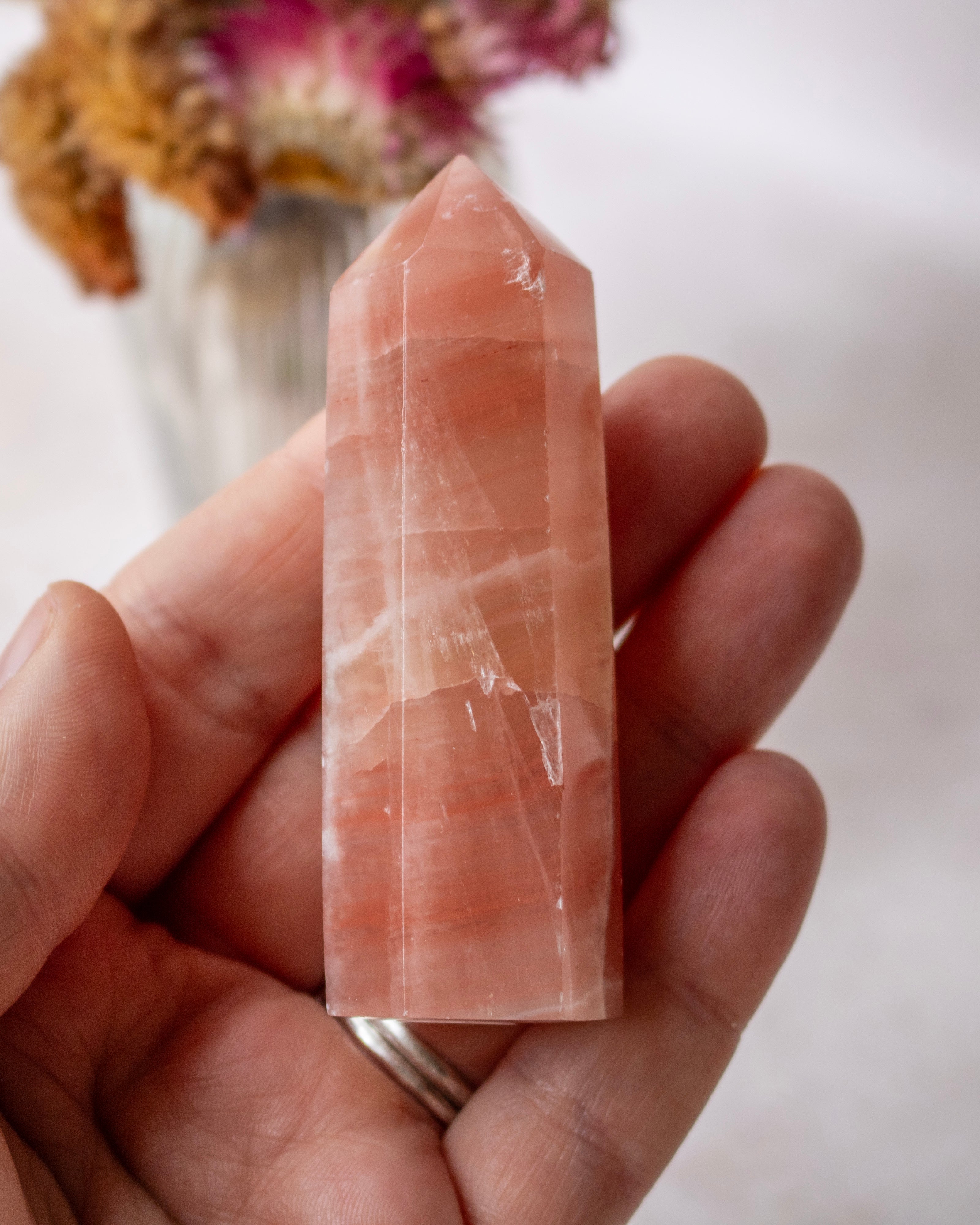 A hand holding a polished rose quartz crystal point against a soft, light background with blurred dried flowers in the distance.