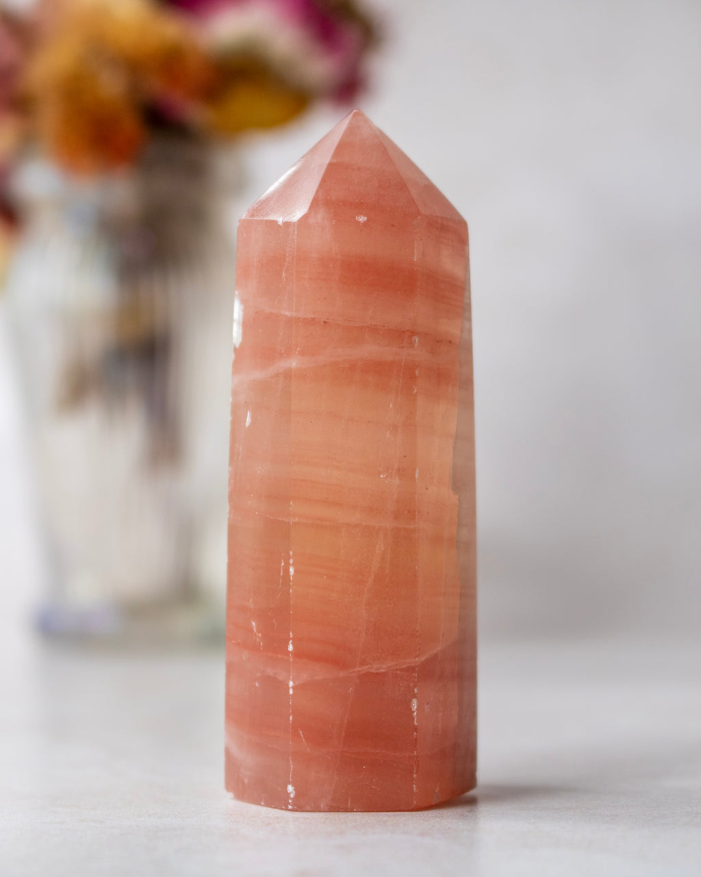 A polished orange-pink crystal point tower, likely made of rose quartz or calcite, standing on a light surface with a softly blurred background.