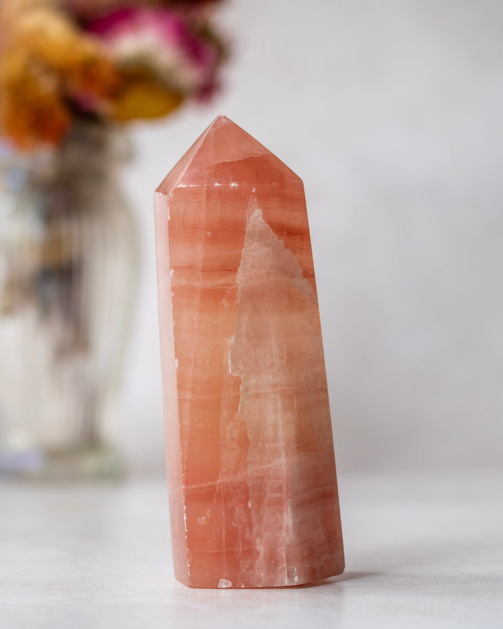 A polished peach-orange quartz point crystal standing upright on a light surface, with a softly blurred vase of flowers in the background.