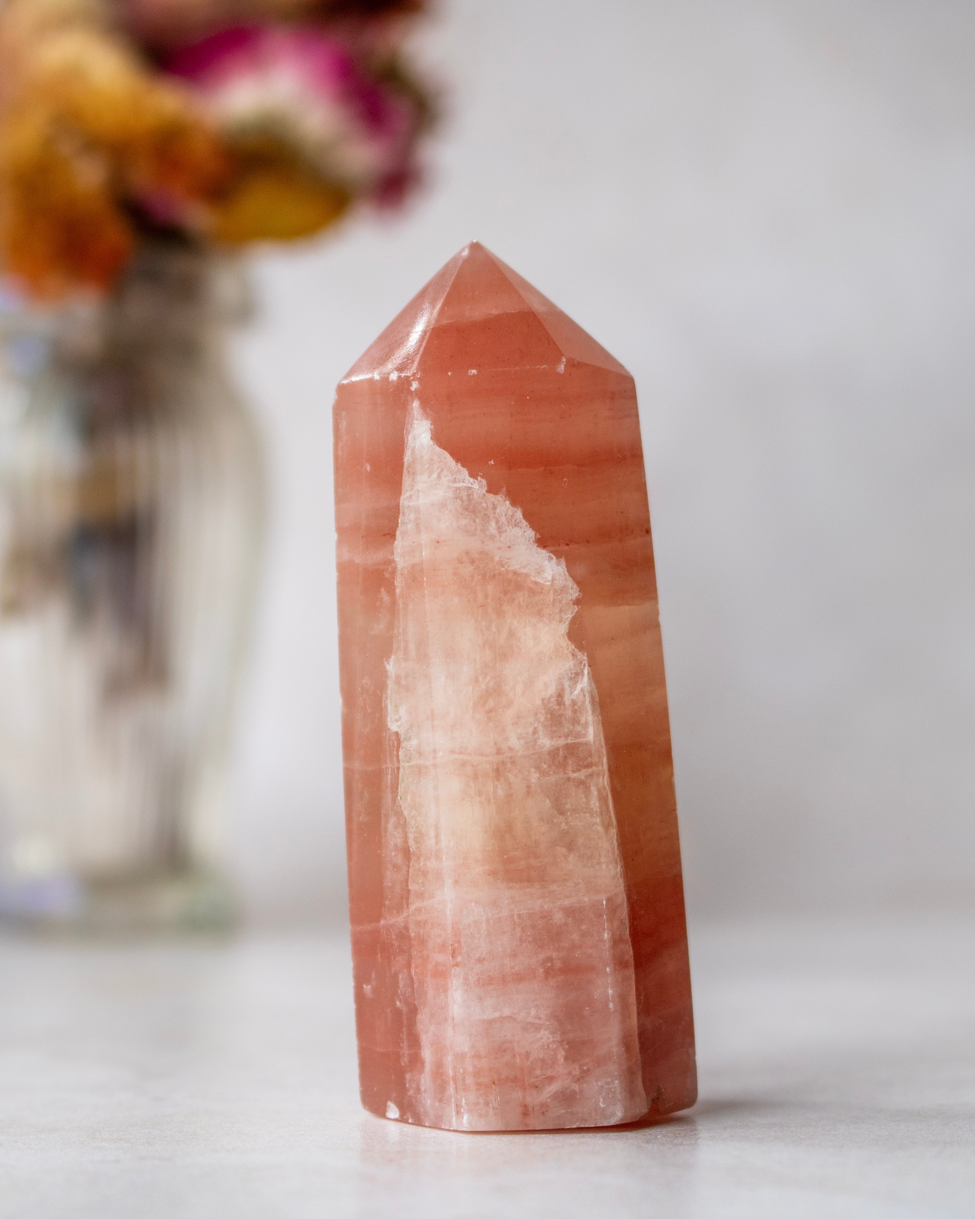 A natural rose calcite crystal point with distinctive orange-pink and white banding, standing on a light surface with a blurred vase of flowers in the background.