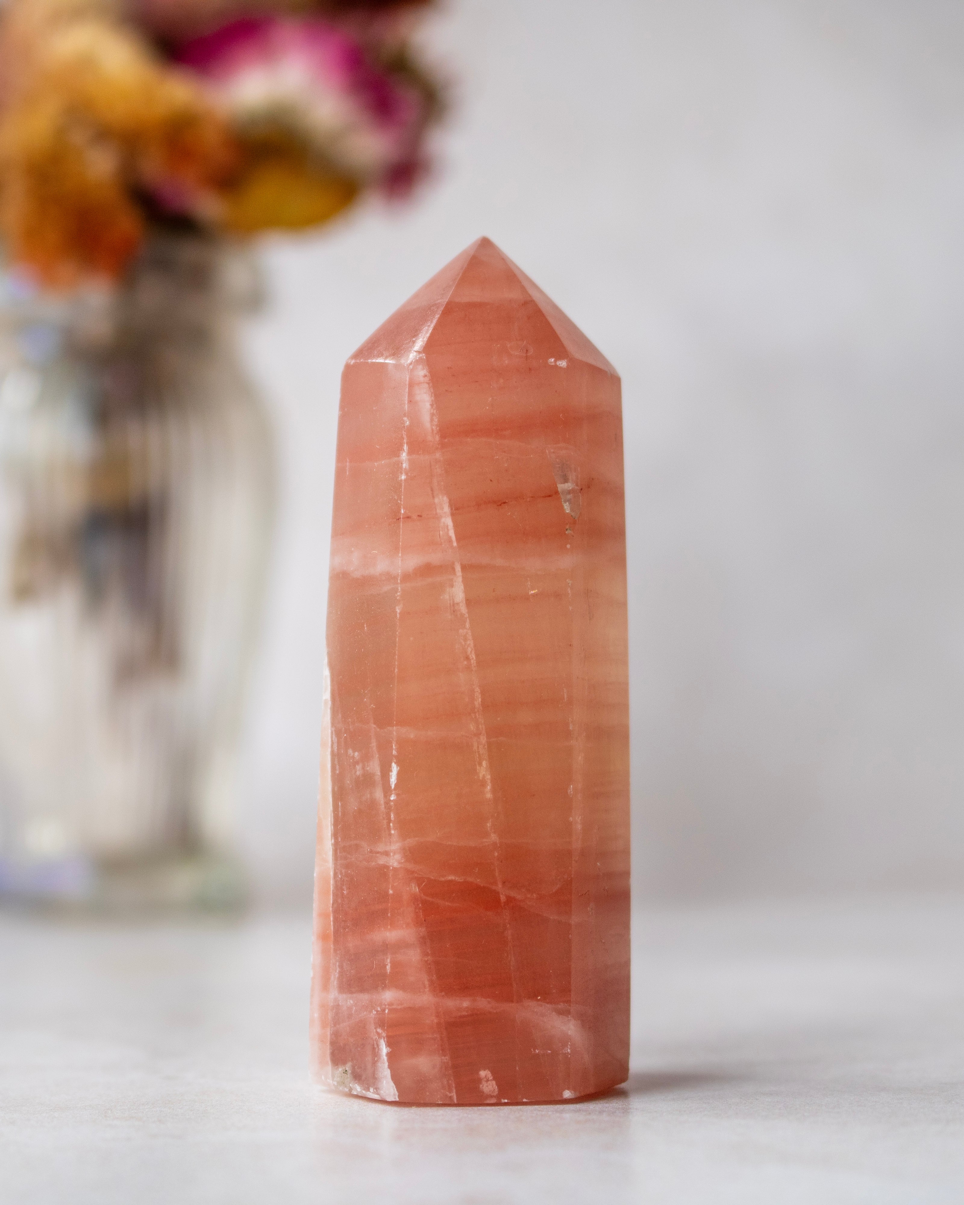 A polished orange-pink crystal point tower with natural banding, standing on a light marble surface with a blurred glass vase of dried flowers in the background.