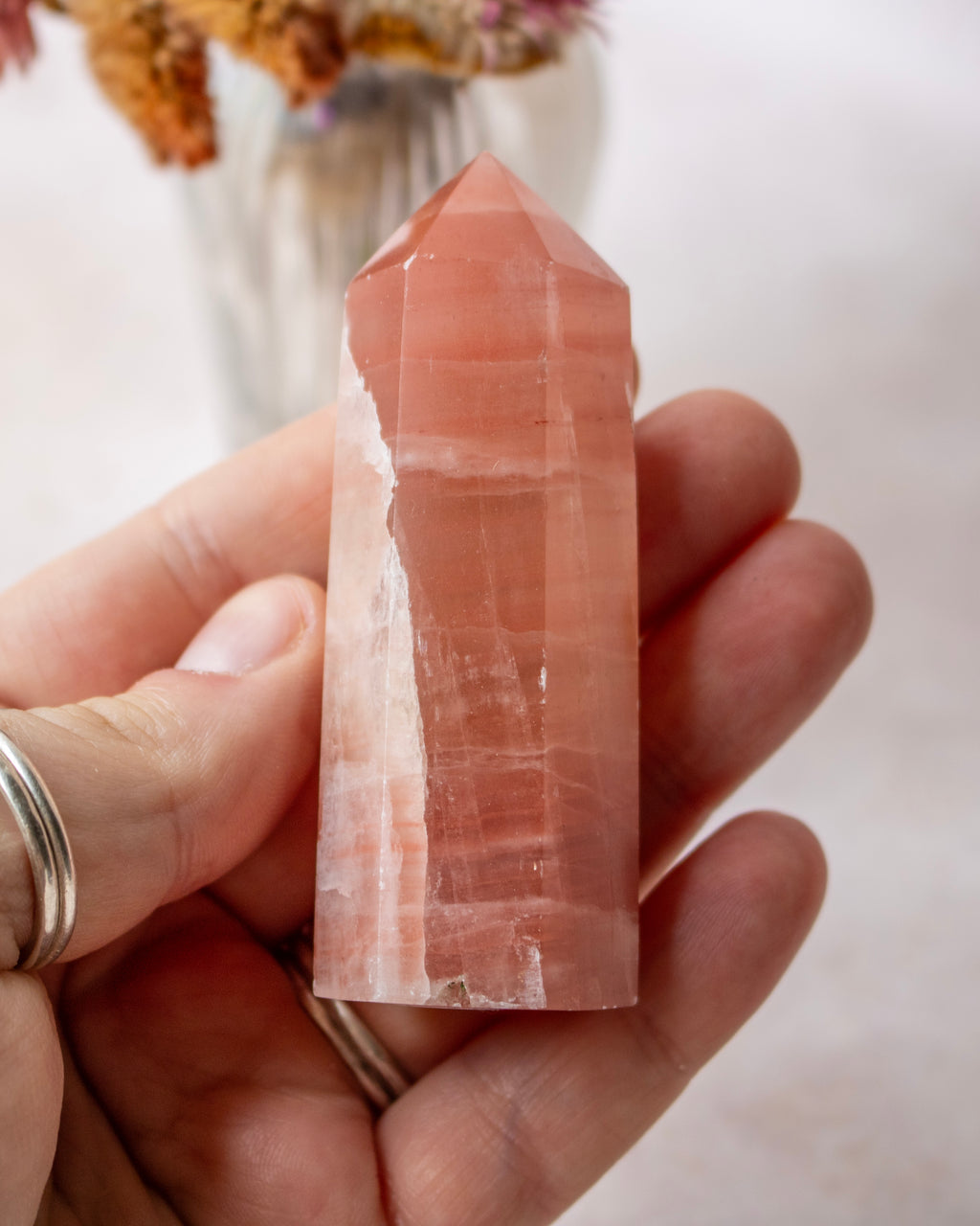 A hand holding a tall, polished rose calcite crystal point against a soft, light background with blurred dried flowers in the distance.