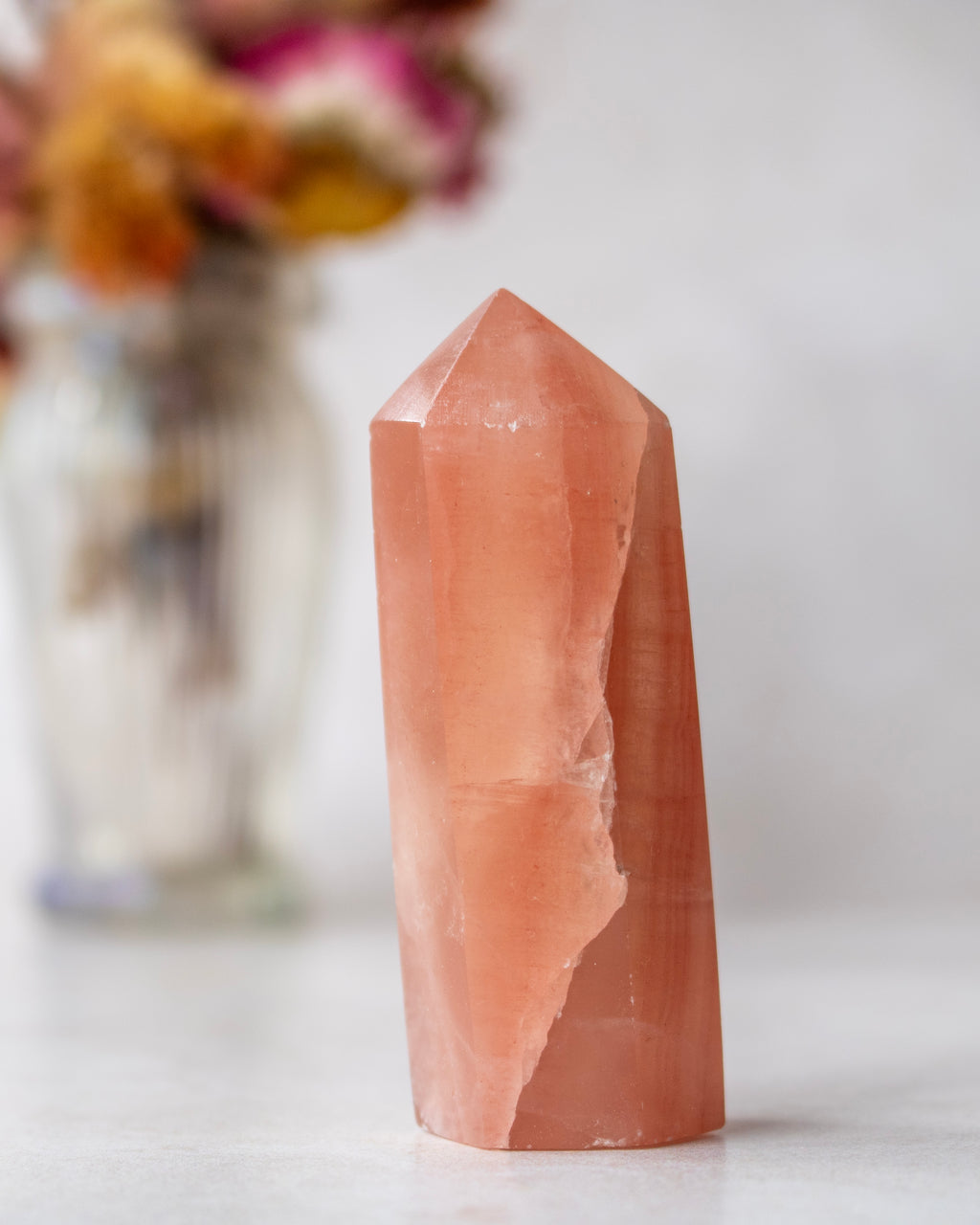 A natural rose quartz crystal point standing upright on a light surface, with a blurred glass vase of dried flowers in the background.