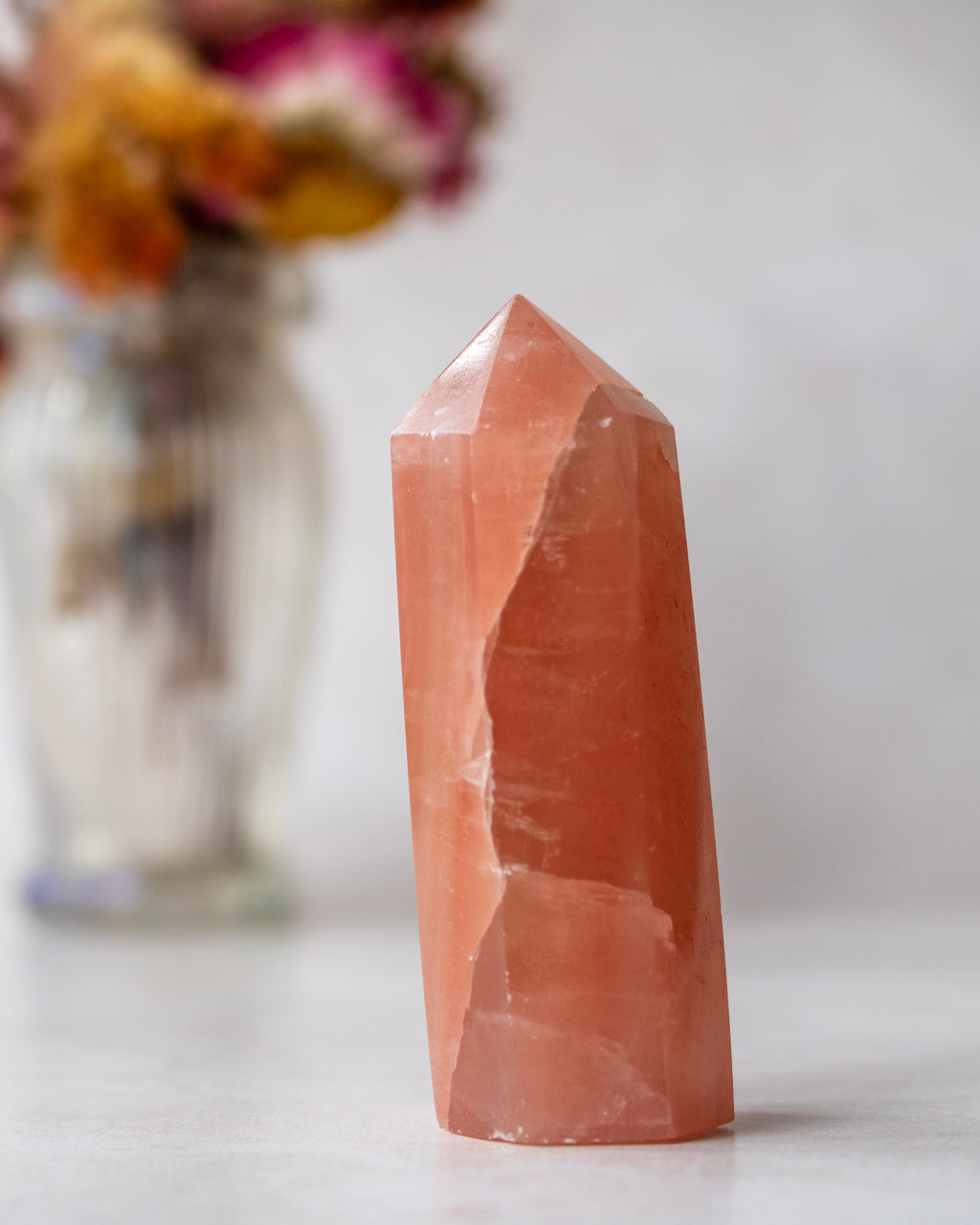 A natural pointed quartz crystal tower in a warm peach-orange hue, standing upright on a light surface with a blurred glass vase of dried flowers in the background.