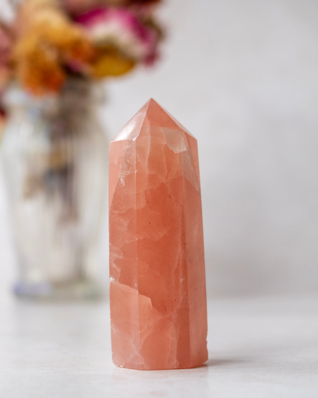 A polished rose quartz crystal tower standing upright on a light surface, with a softly blurred glass vase of dried flowers in the background.